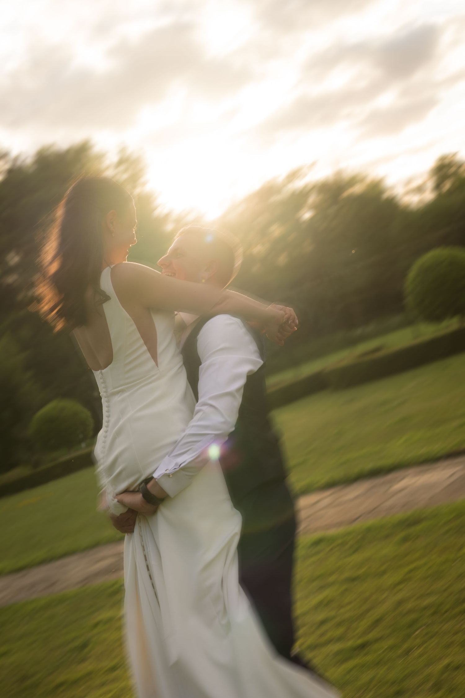 A bride and groom share a romantic spin at sunset in a lush garden, with golden light and lens flare.