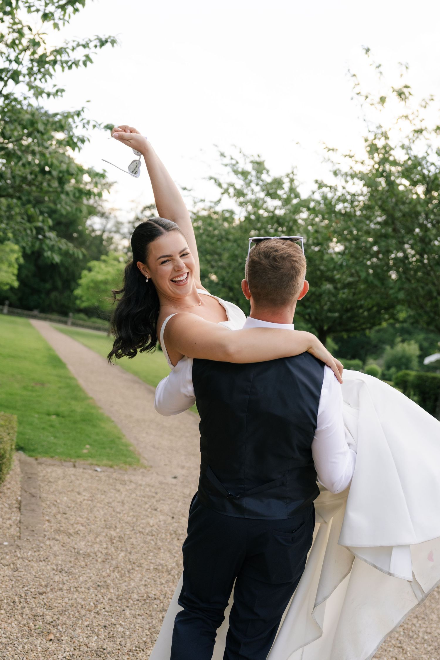 Groom carrying bride in white dress outdoors, bride laughing with arm raised, lush green park pathway background.