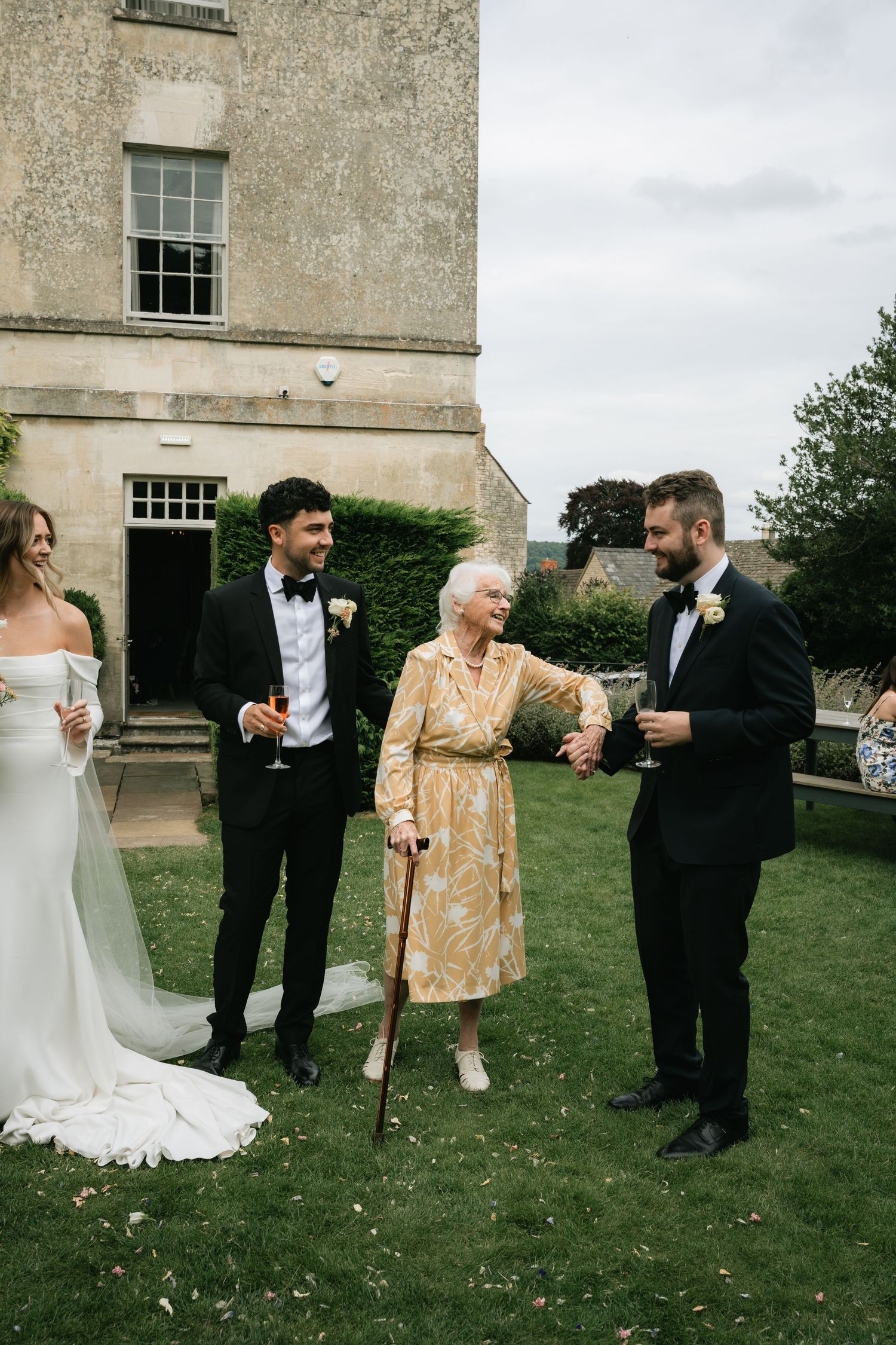 A wedding celebration on a mansion lawn with guests in formal attire including a gold dress and black tuxedos.