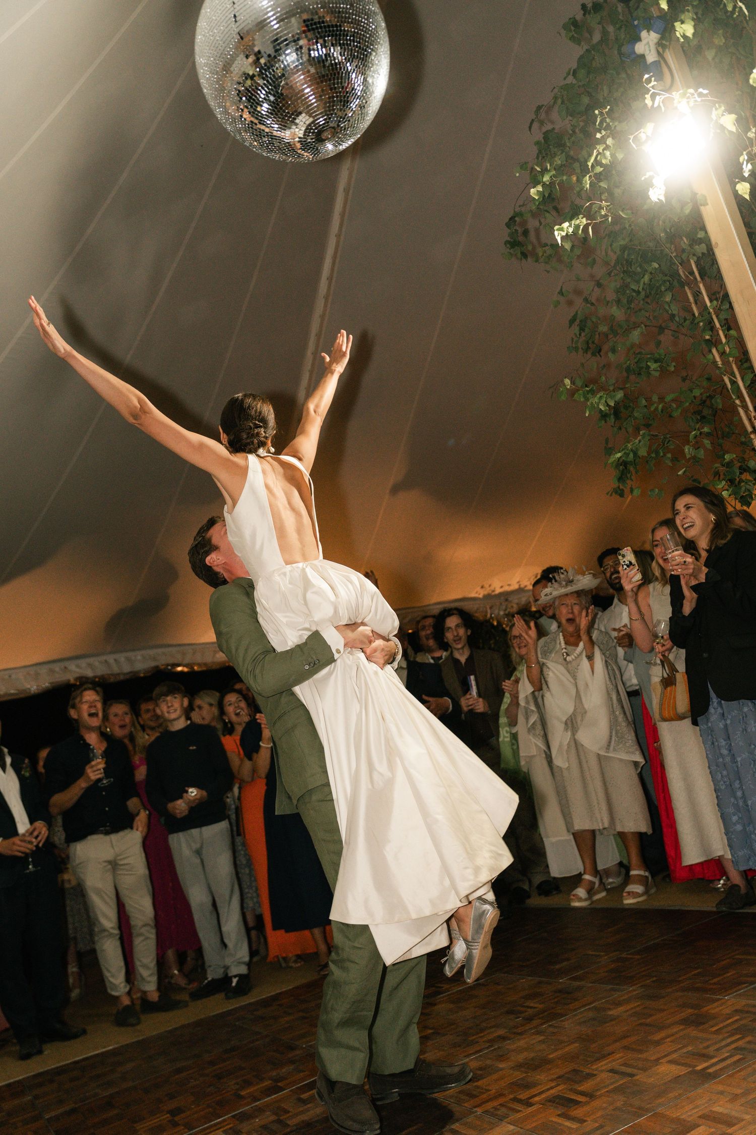 A couple performs a dramatic dance lift under a disco ball at a wedding reception on a wooden dance floor.