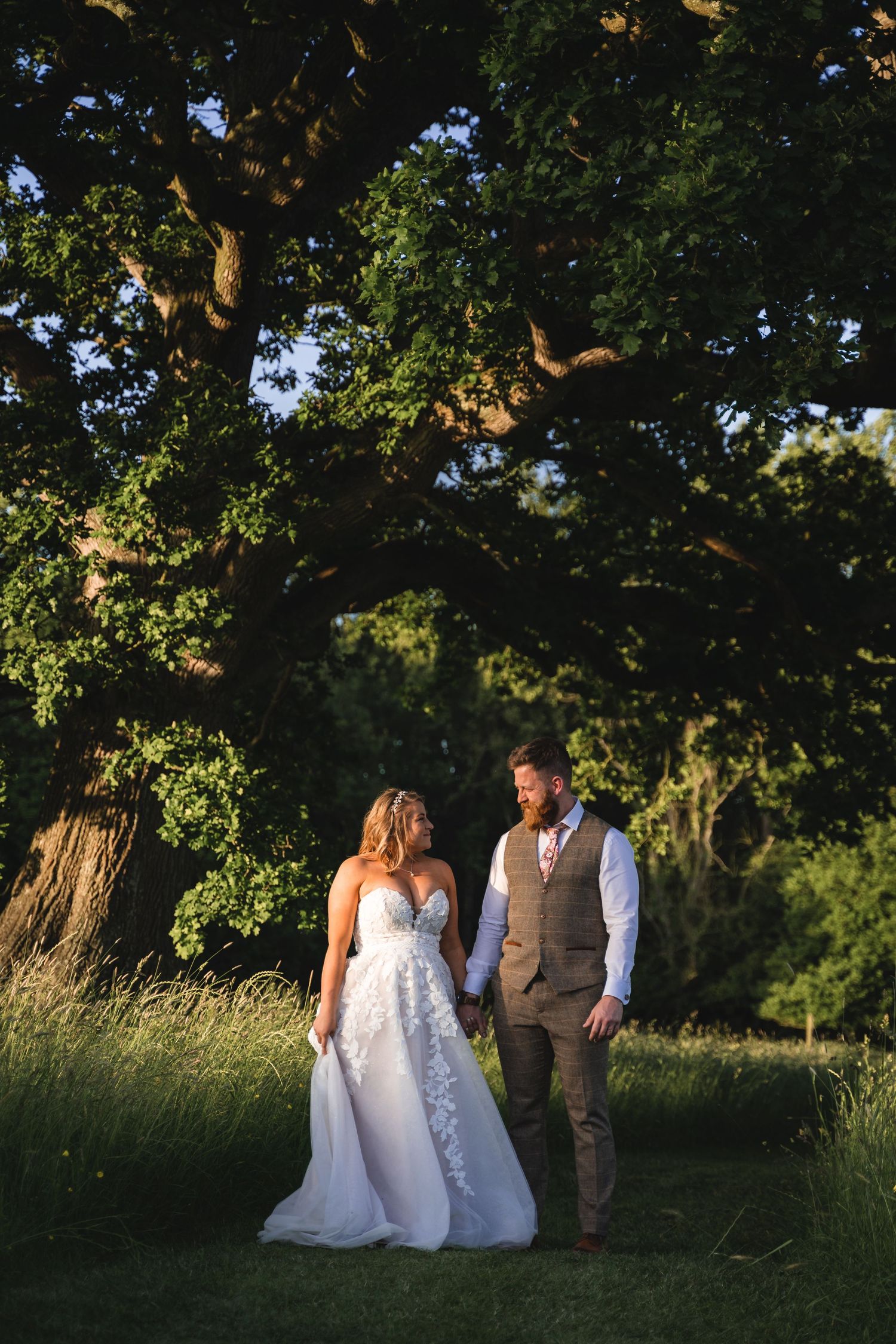 Wedding couple walks through a scenic meadow at sunset with trees in background.