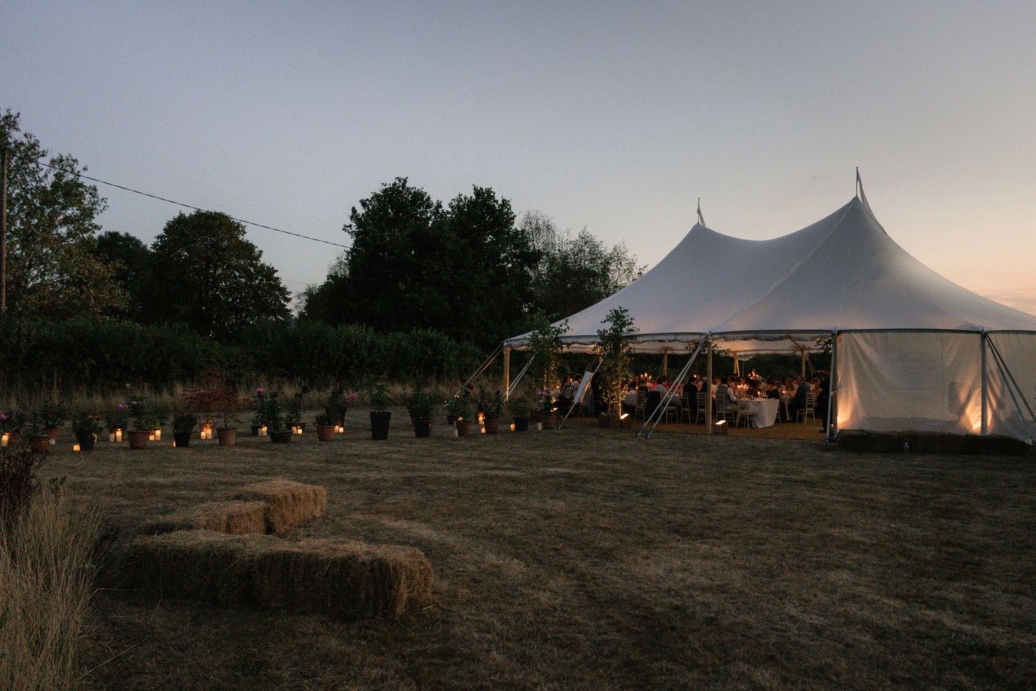 A series of white event tents set up on a grassy hillside at sunset with string lights creating an elegant outdoor atmosphere.