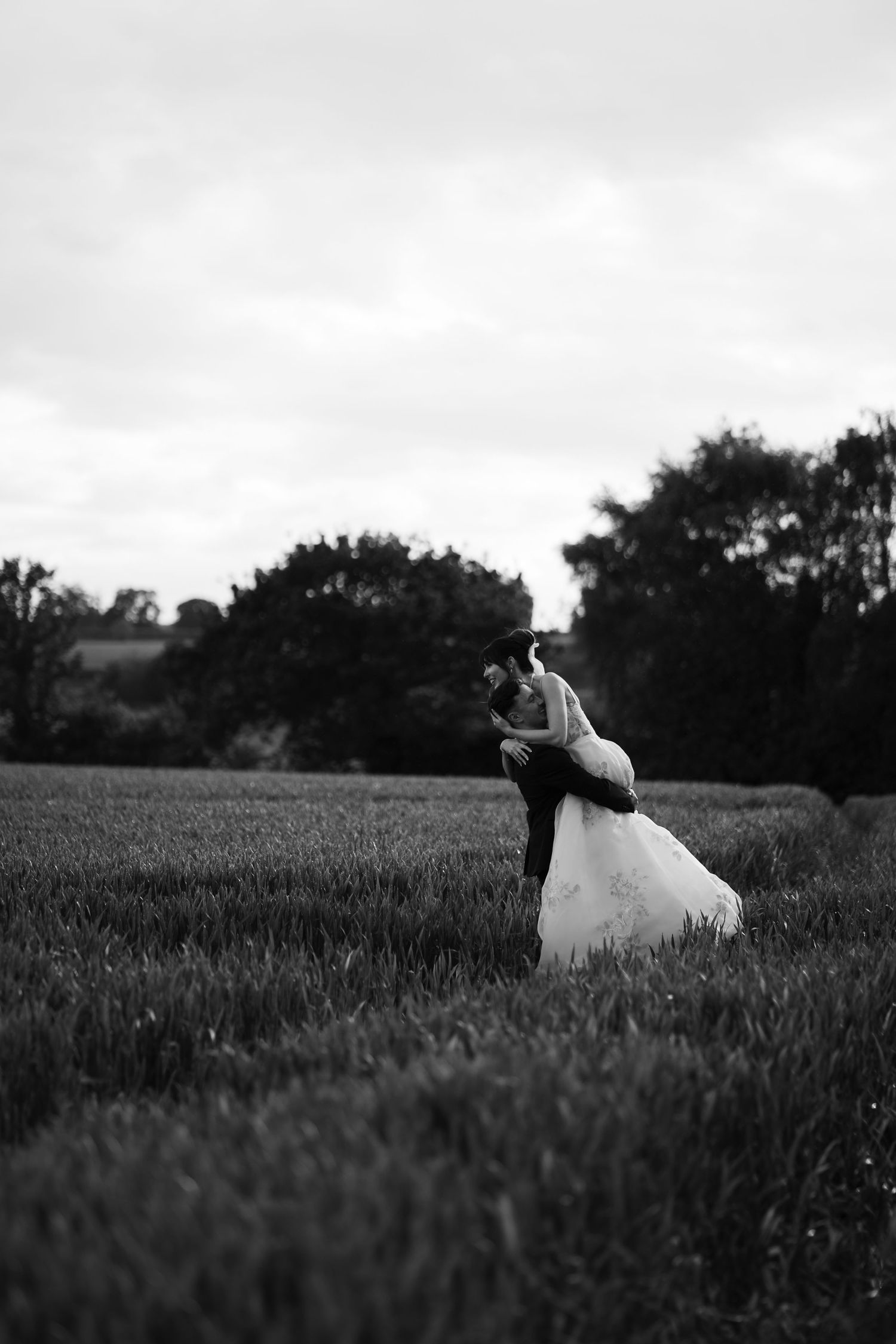 Black and white panoramic photo of a couple in wedding attire embracing in a vast field at dusk.