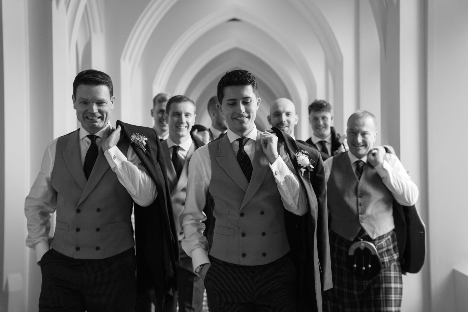 A vintage black and white photograph shows a line of men in formal suits standing in an elegant hallway.