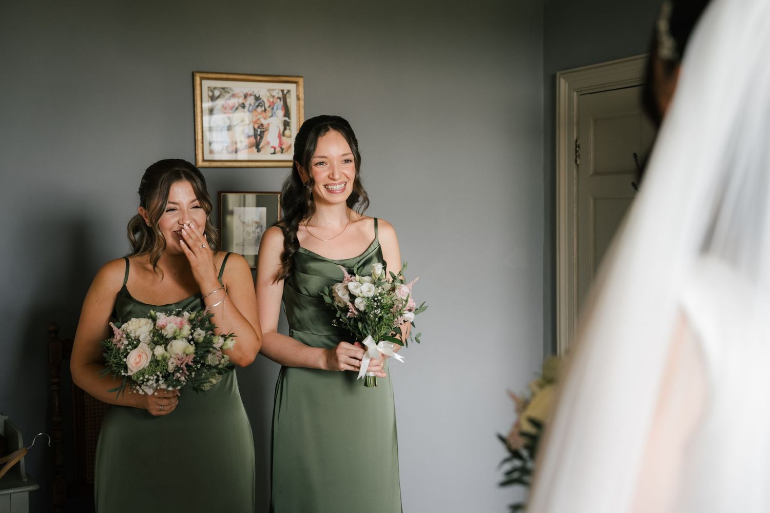 Bridesmaids in sage green dresses help adjust a wedding gown in a softly lit room.
