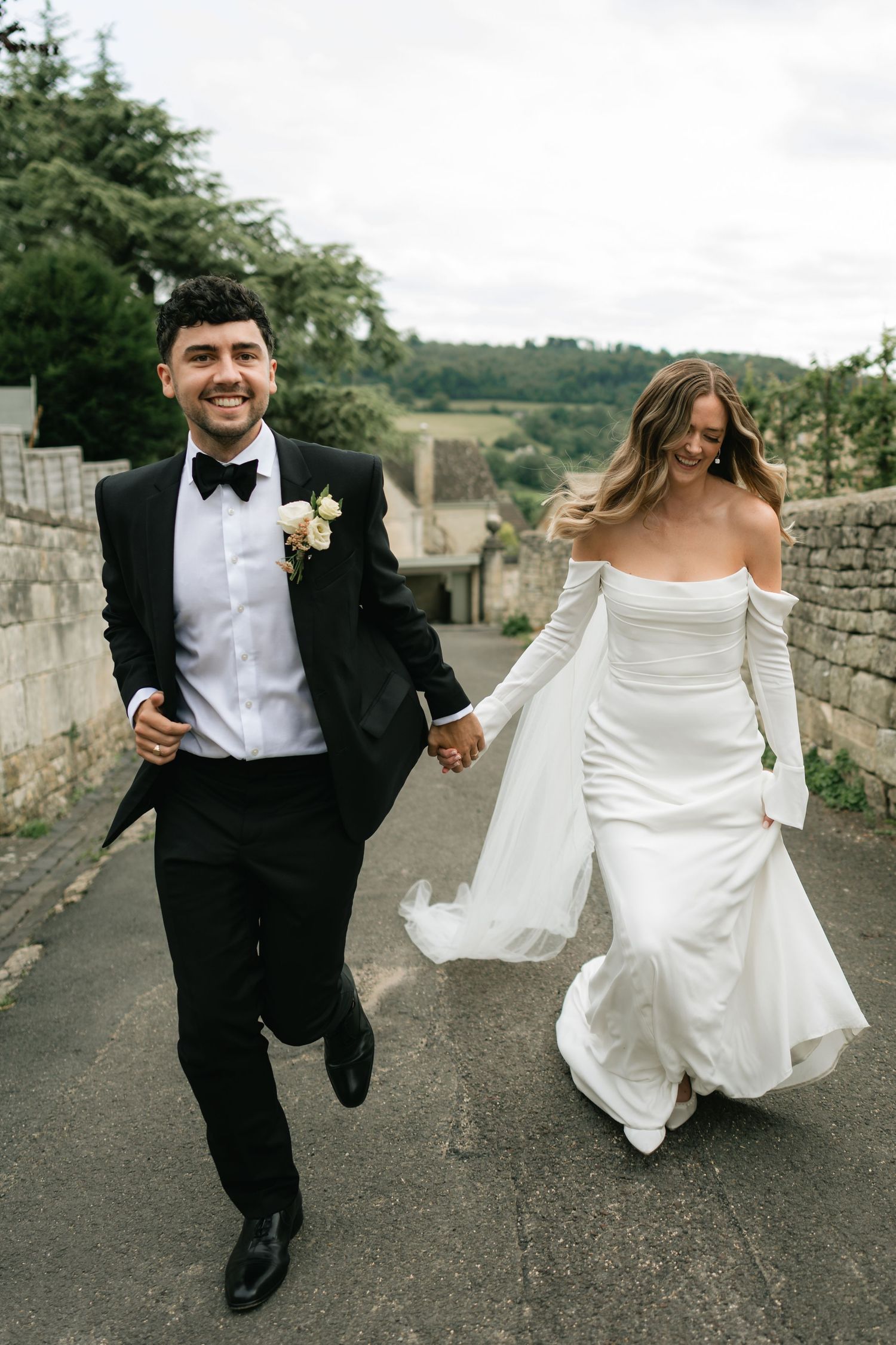 A couple runs playfully along a stone wall path during their wedding celebration.