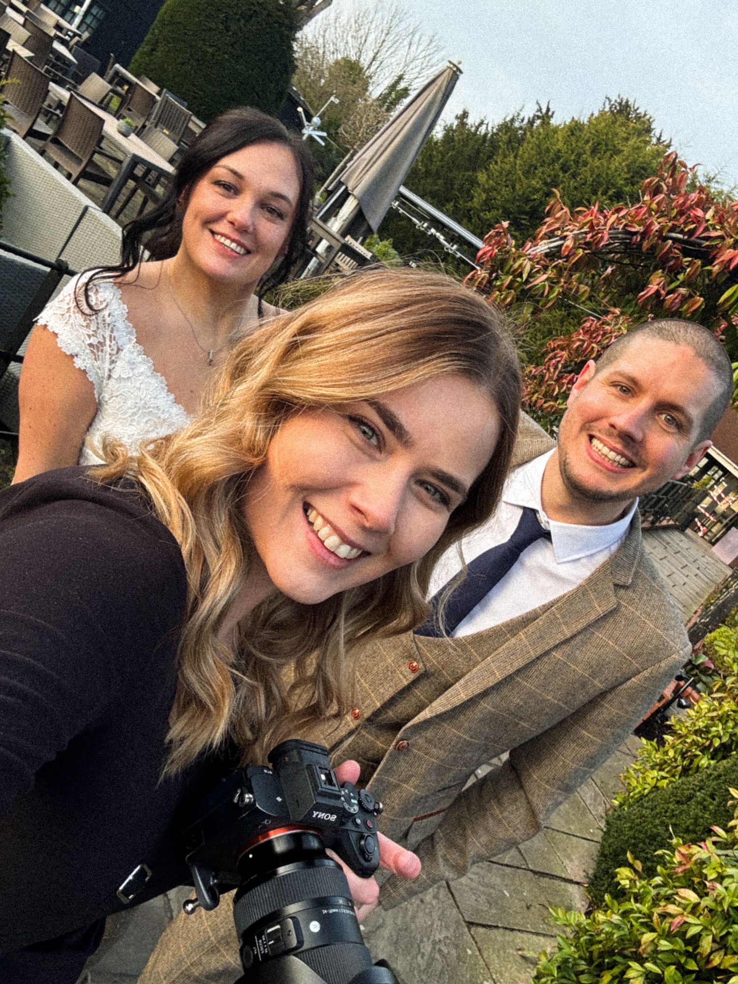 A photographer with camera equipment captures a selfie with others at an outdoor venue with greenery in the background.