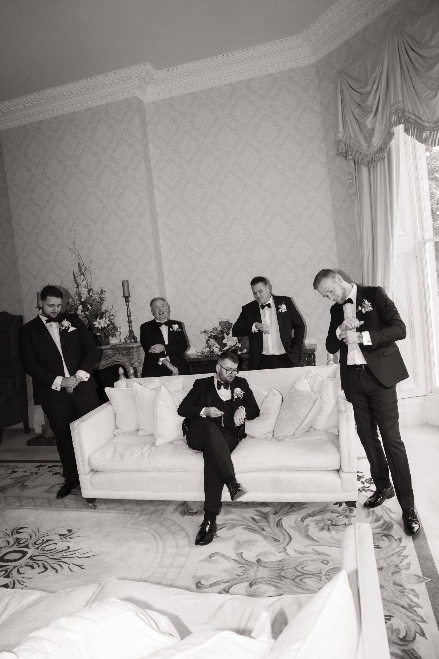 A black and white photo of groomsmen in tuxedos relaxing on and around an elegant white couch in a formal room.