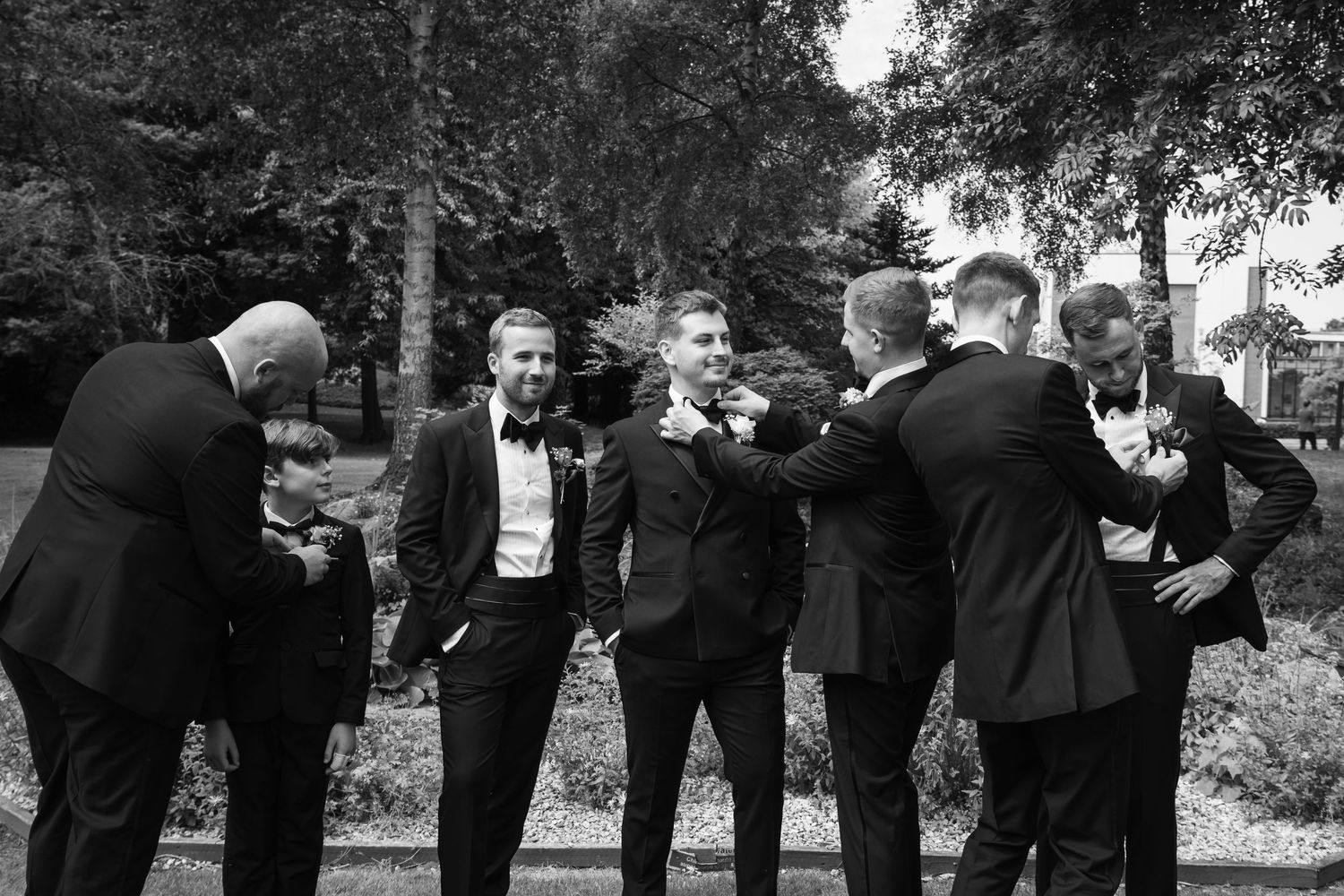 A group of formally dressed men in tuxedos share a candid moment and laughter outdoors in black and white photography.
