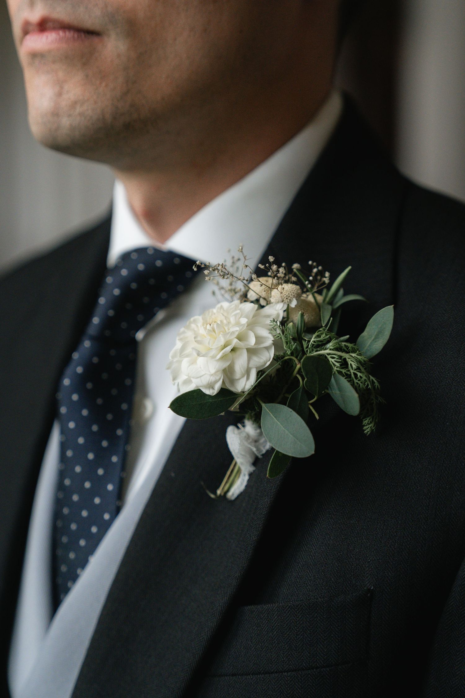 White floral boutonniere with eucalyptus leaves adorns a black suit jacket with polka dot tie.