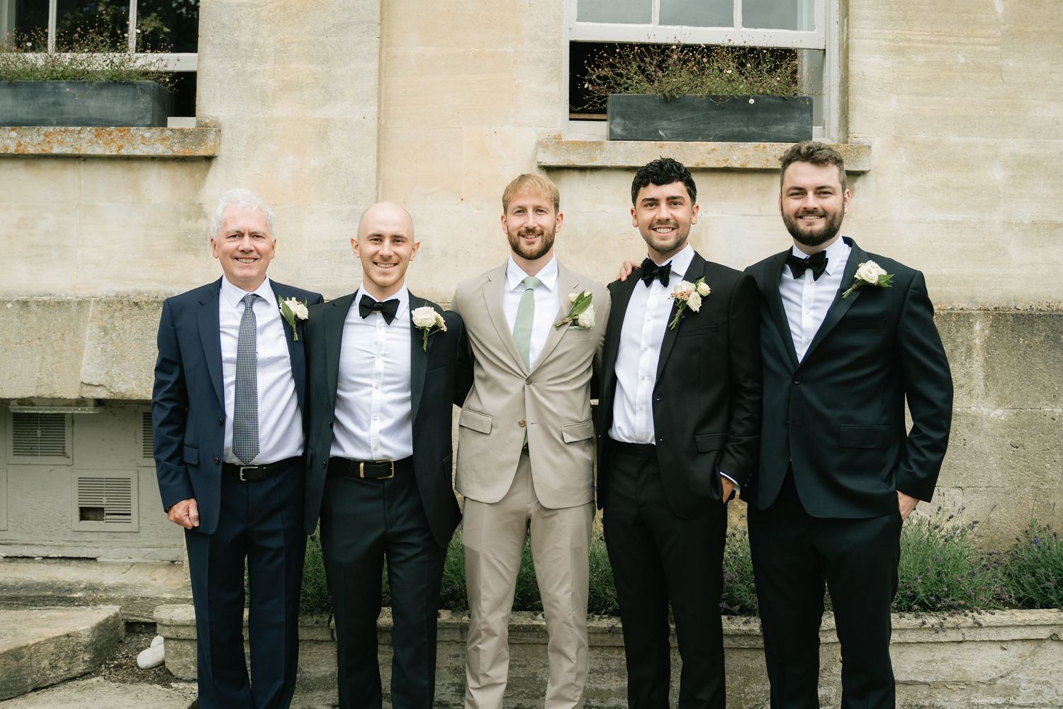 Five men in formal attire stand together smiling outside a stone building for a wedding photo.