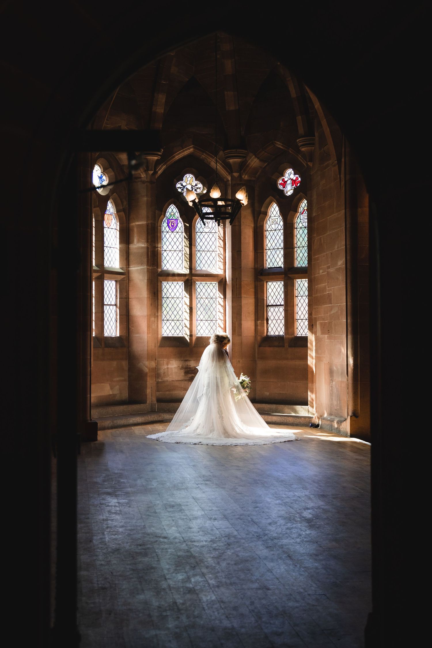 A bride in a white wedding dress stands silhouetted against gothic church windows in a stone chapel.