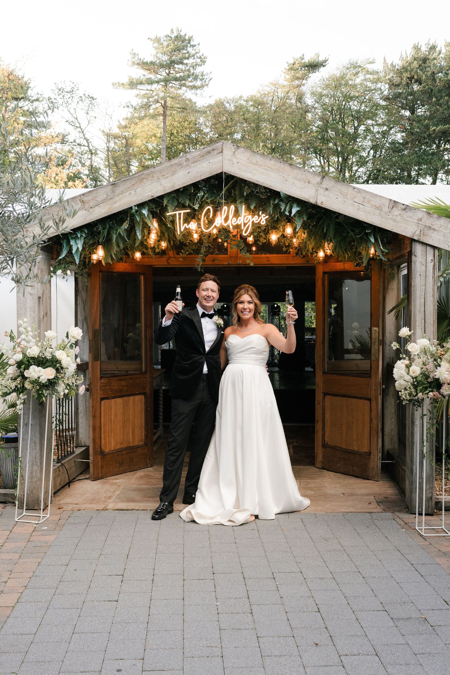 Wedding couple celebrates with guests under rustic wooden pavilion decorated with string lights and floral arrangements.