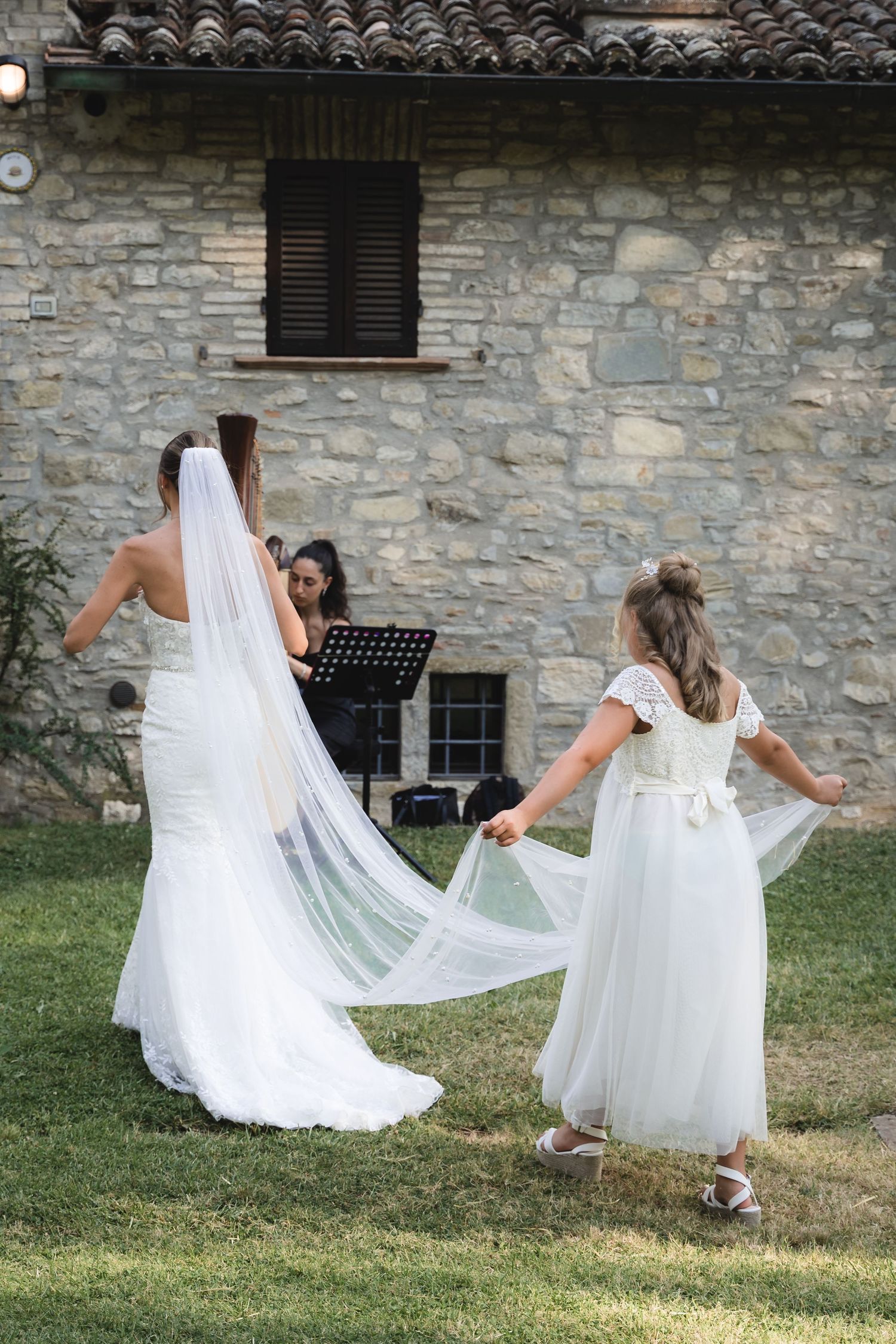 A bride in a flowing white dress and veil walks with a flower girl in a matching white dress on a grassy lawn.