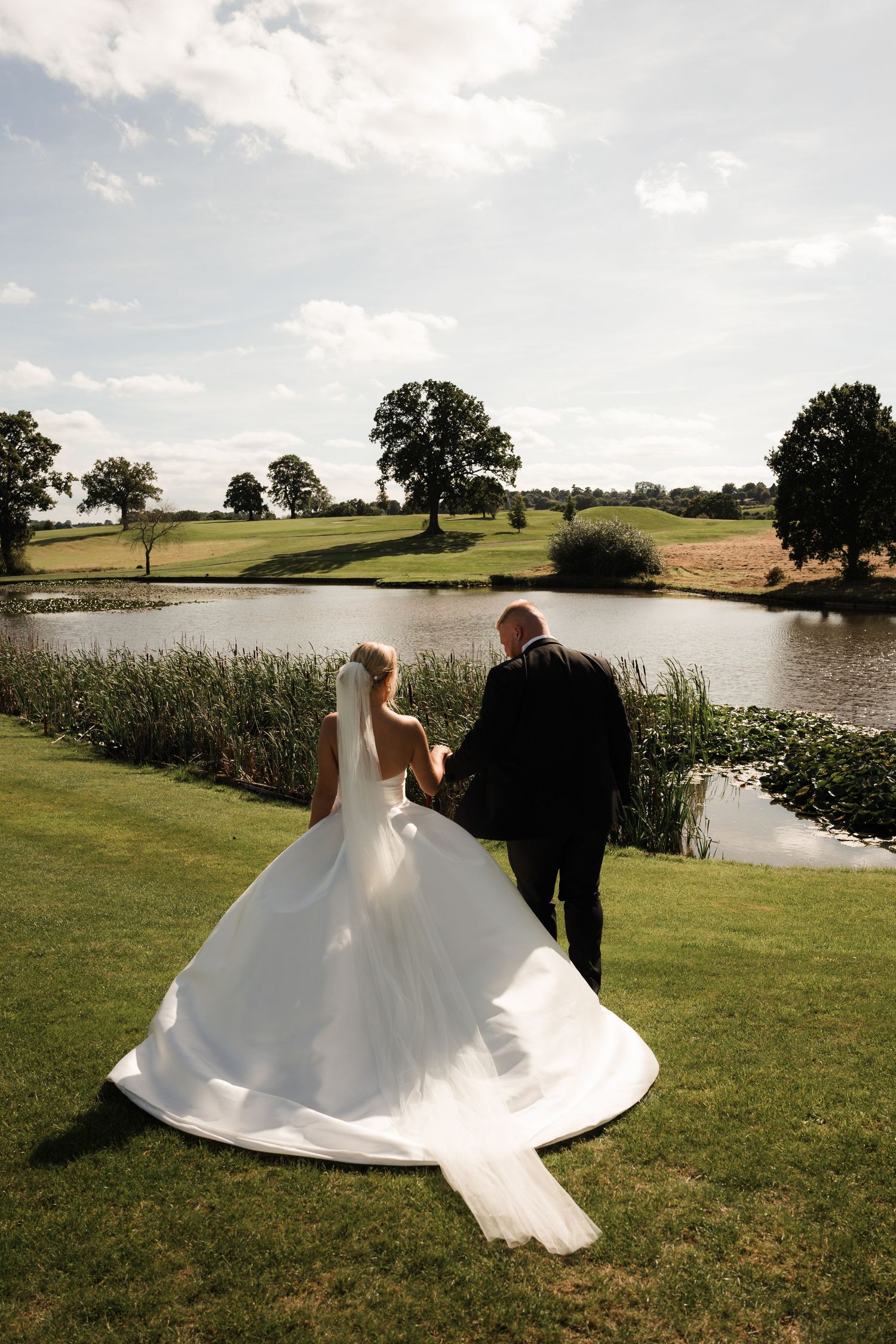 A wedding couple twirls on lush green grass near a scenic pond, making the bride's white dress flow dramatically.