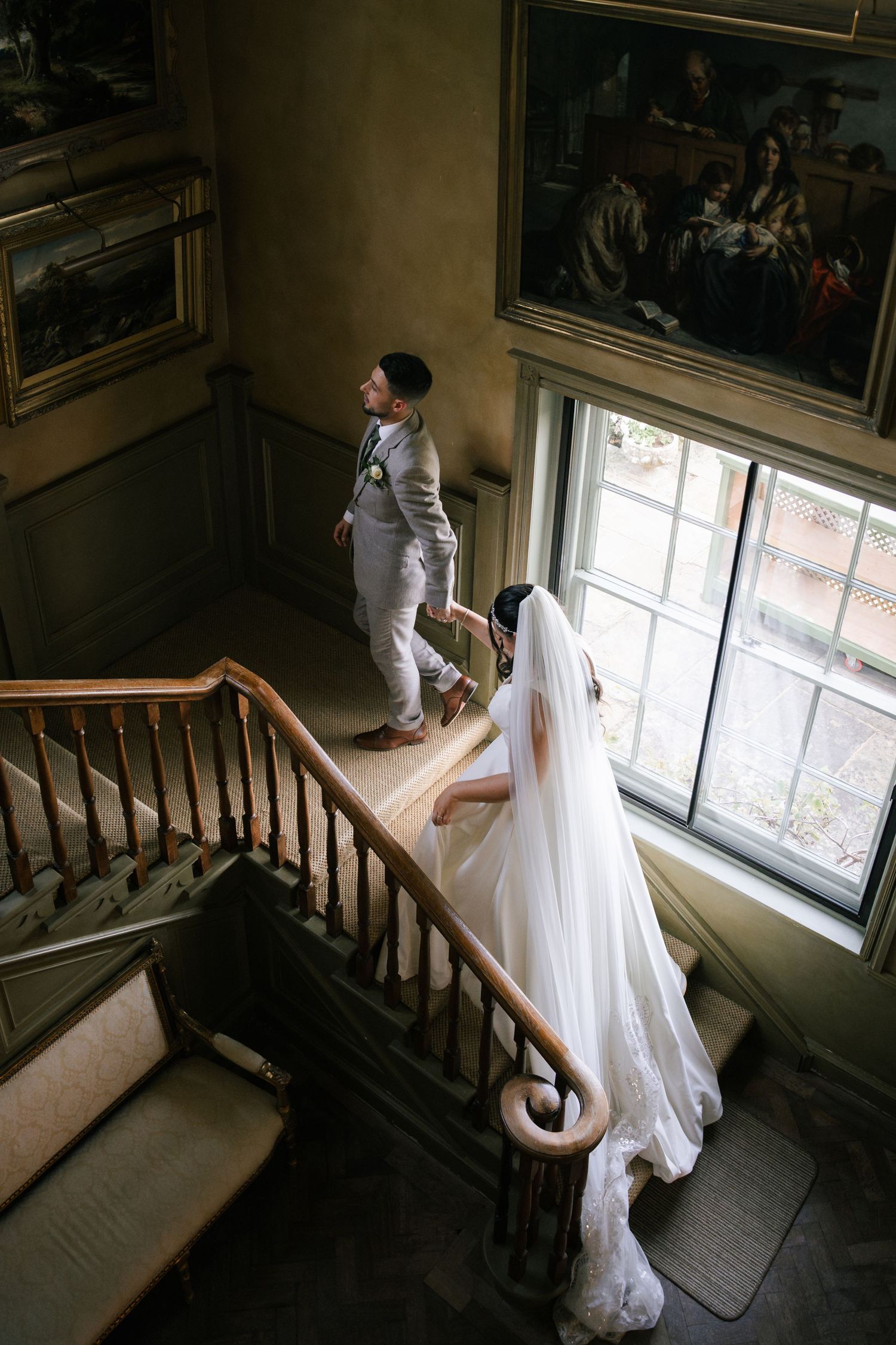 A bride in a flowing white wedding dress stands by a window on a wooden staircase in a historic building.