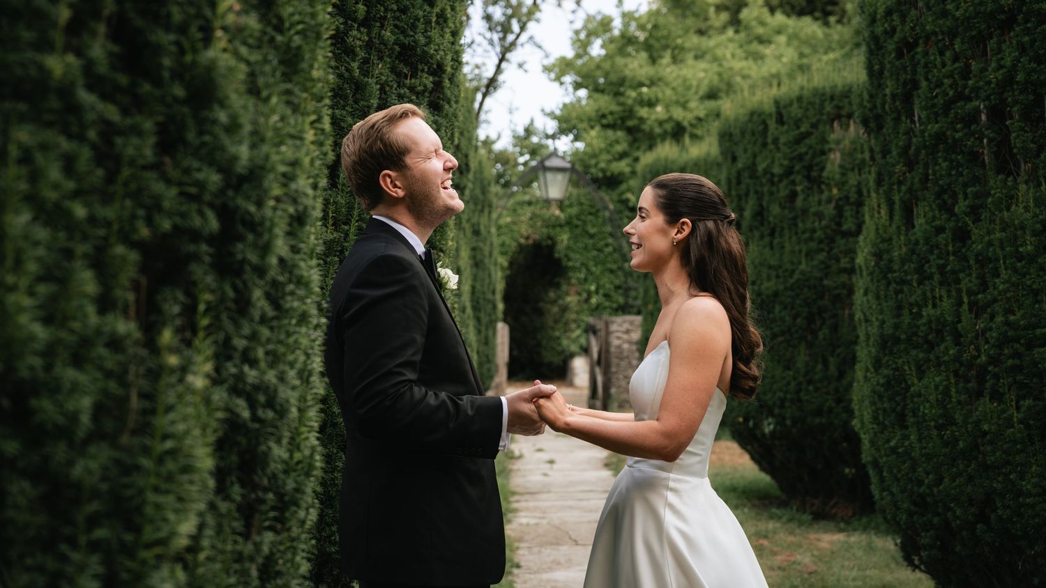 A bride and groom hold hands and smile at each other in a lush green garden on their wedding day.