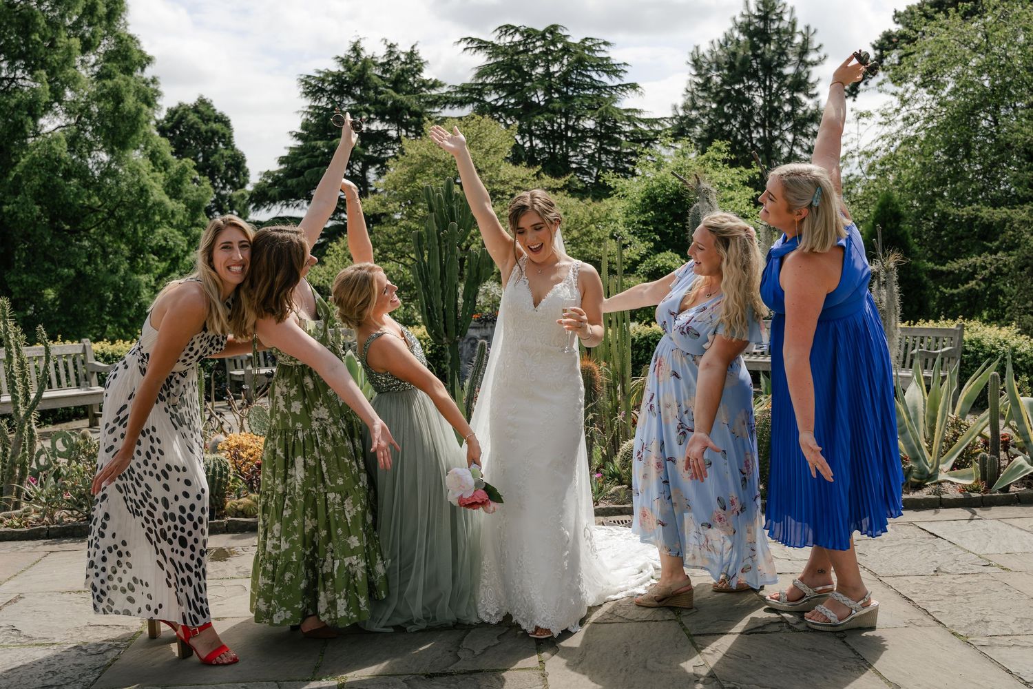 A celebratory group in colorful dresses raise their arms joyfully on a stone patio surrounded by lush garden scenery.