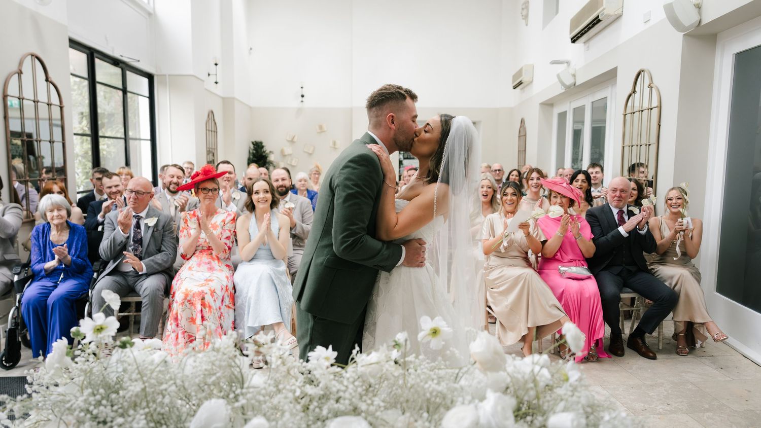 A romantic wedding ceremony in a bright white venue with baby's breath floral decorations and seated guests watching.