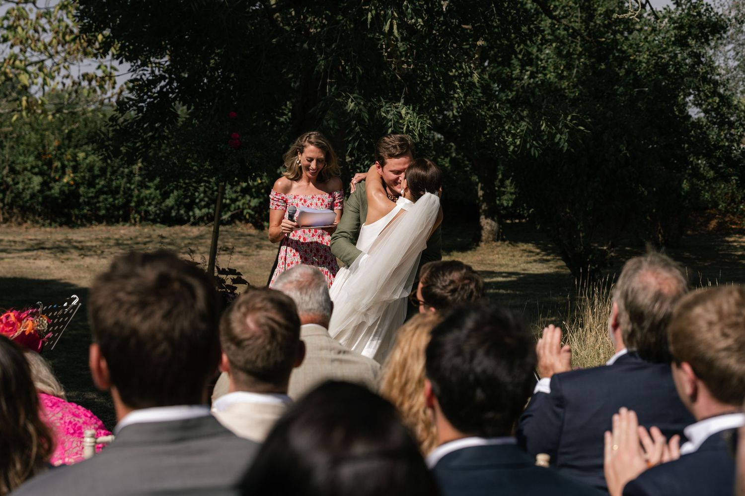 A wedding ceremony takes place outdoors with guests seated in white chairs under trees during a sunny day.