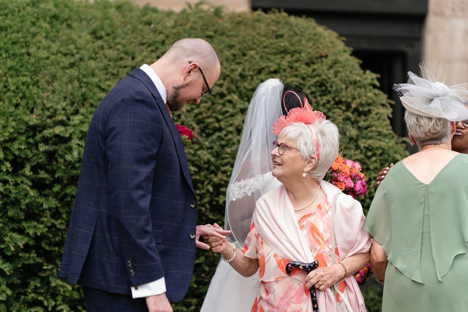 Wedding guests in formal attire interact outdoors near green hedges during a sunny celebration.