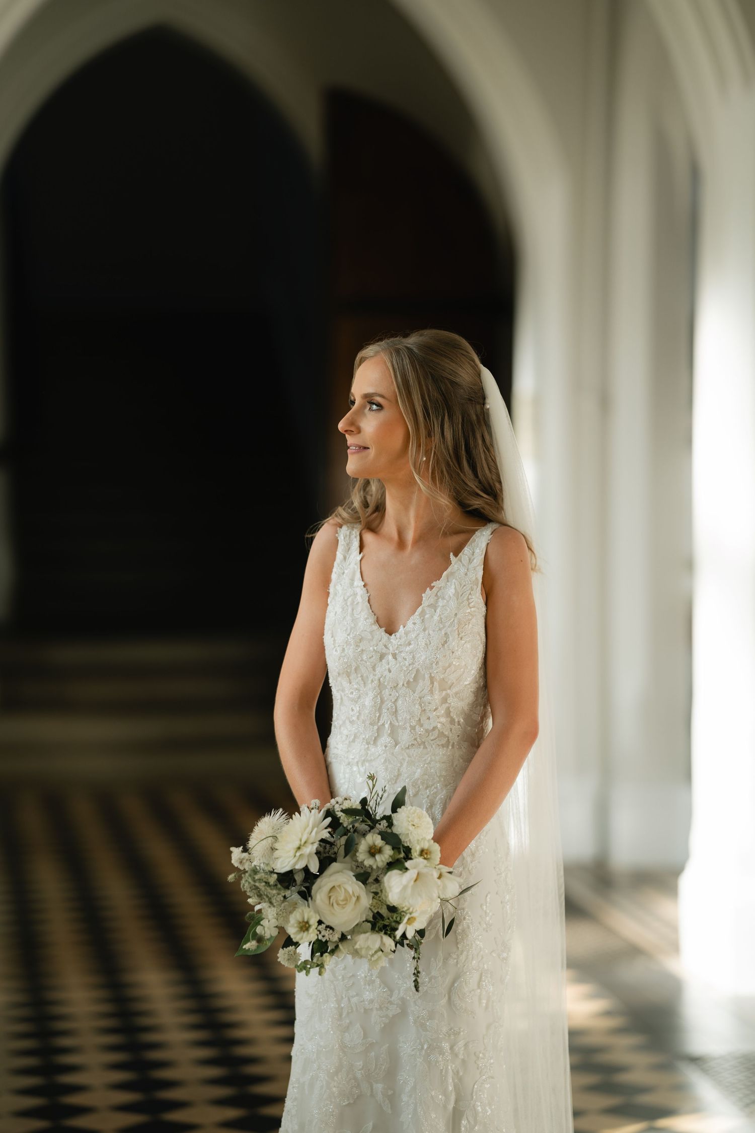 A bride in a white lace wedding dress holds a white and green bouquet in an elegant hallway with checkered floors.