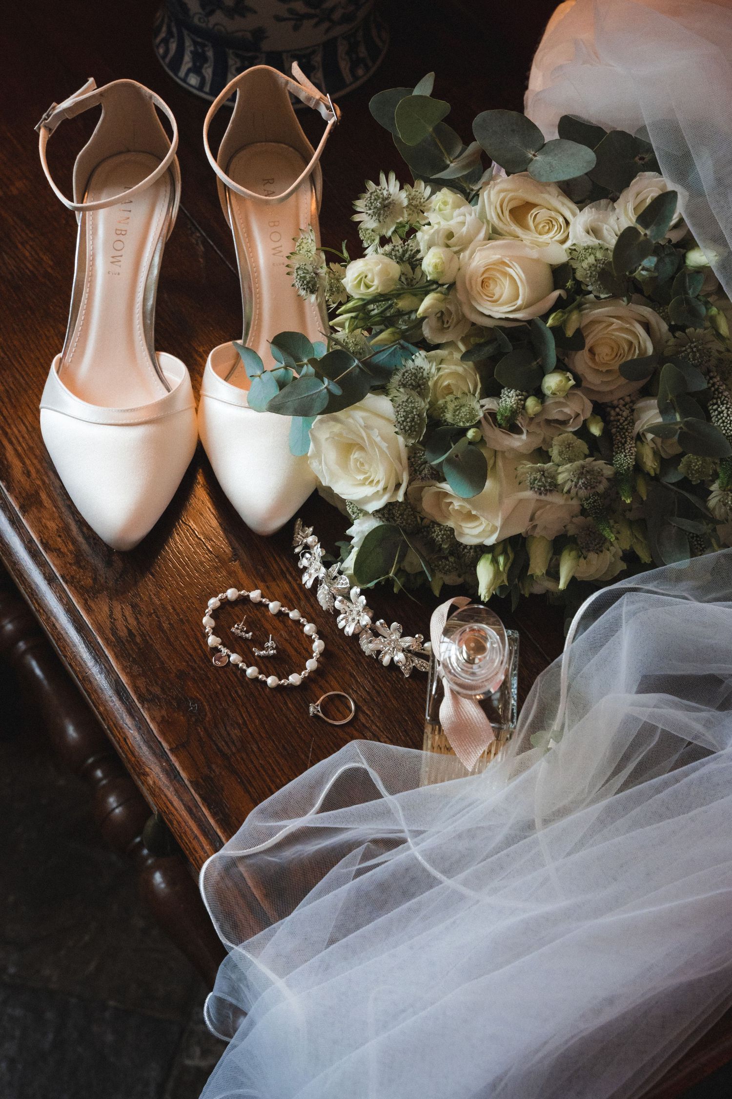 White bridal shoes, veil, and floral bouquet arranged on wooden surface with wedding rings.