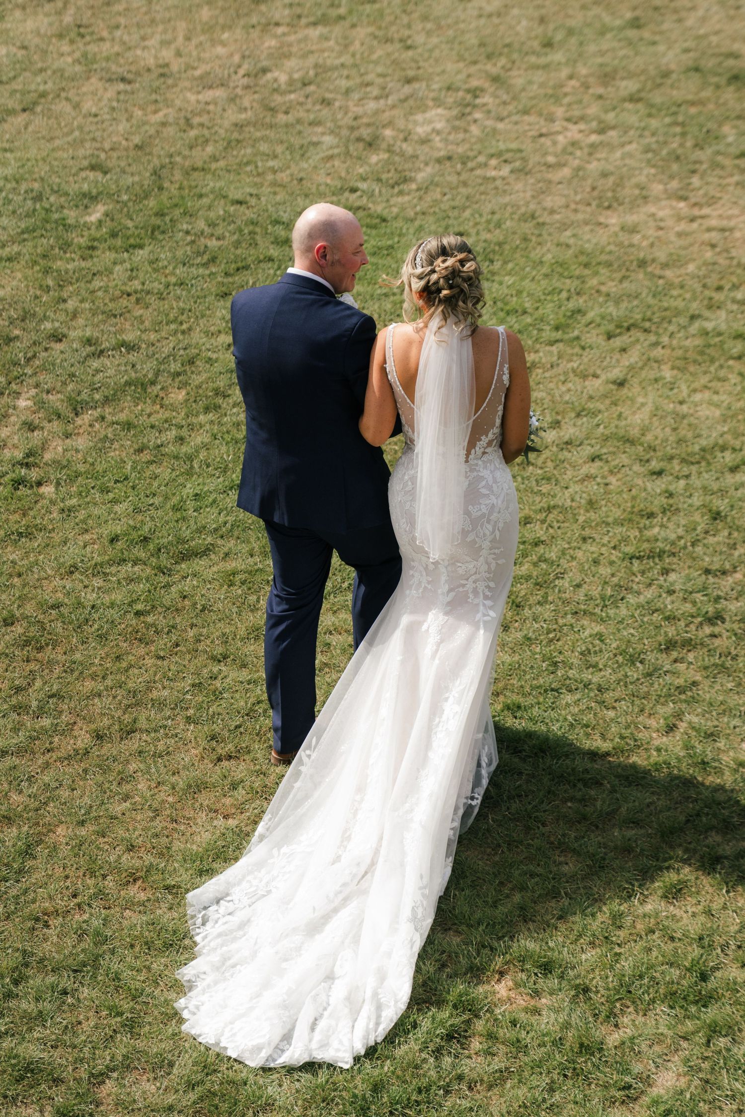 A bride and groom walk together on a grassy lawn, with the bride's white lace dress trailing behind them.