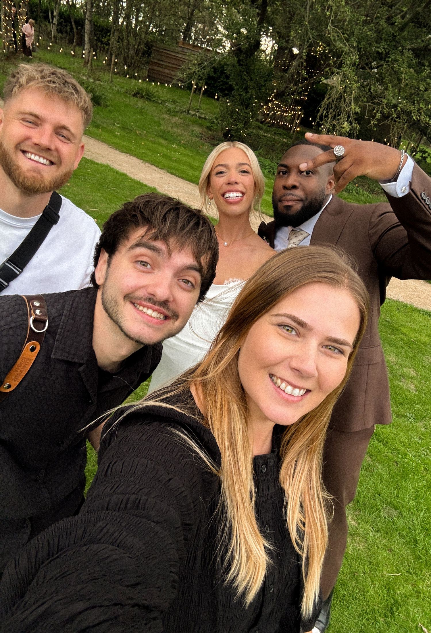 Group of five smiling friends posing for a selfie outdoors at a wedding celebration on a green lawn.