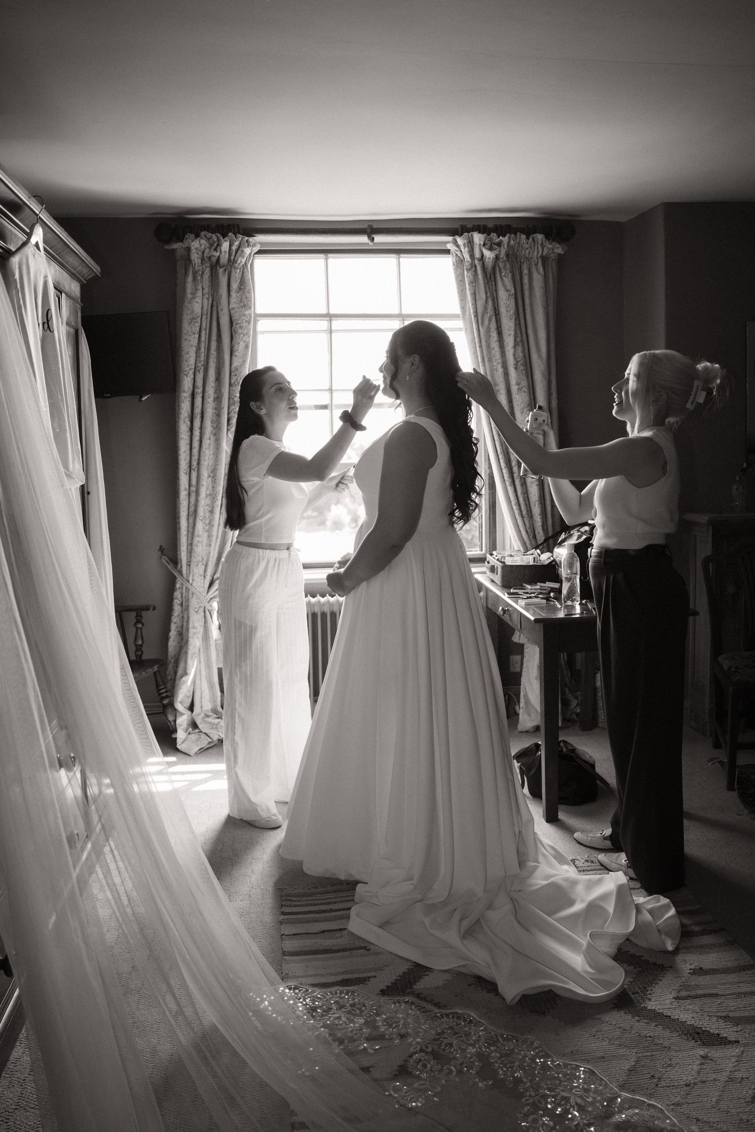 Black and white photo of wedding preparations with a bride in a flowing white dress being helped with final touches.