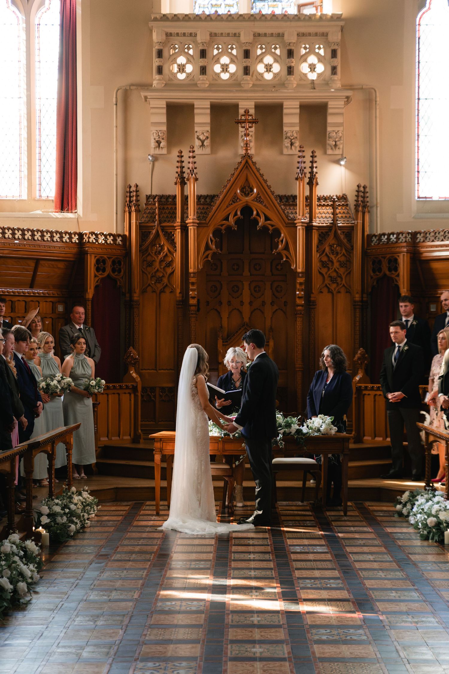 A wedding ceremony takes place in a beautiful historic church with ornate wooden paneling and sunlight streaming through windows.