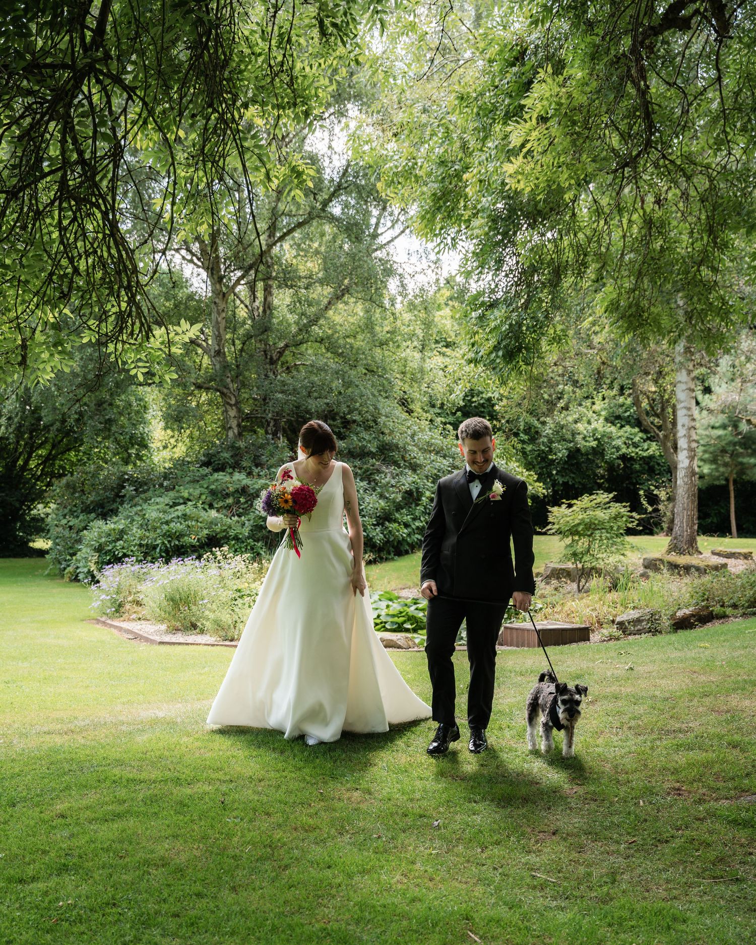 A couple walks through a lush garden setting with their dog during an outdoor wedding photoshoot.