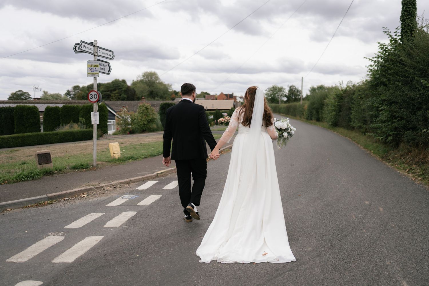 Wedding couple walks along a country road while holding hands in a romantic post-ceremony photo series.