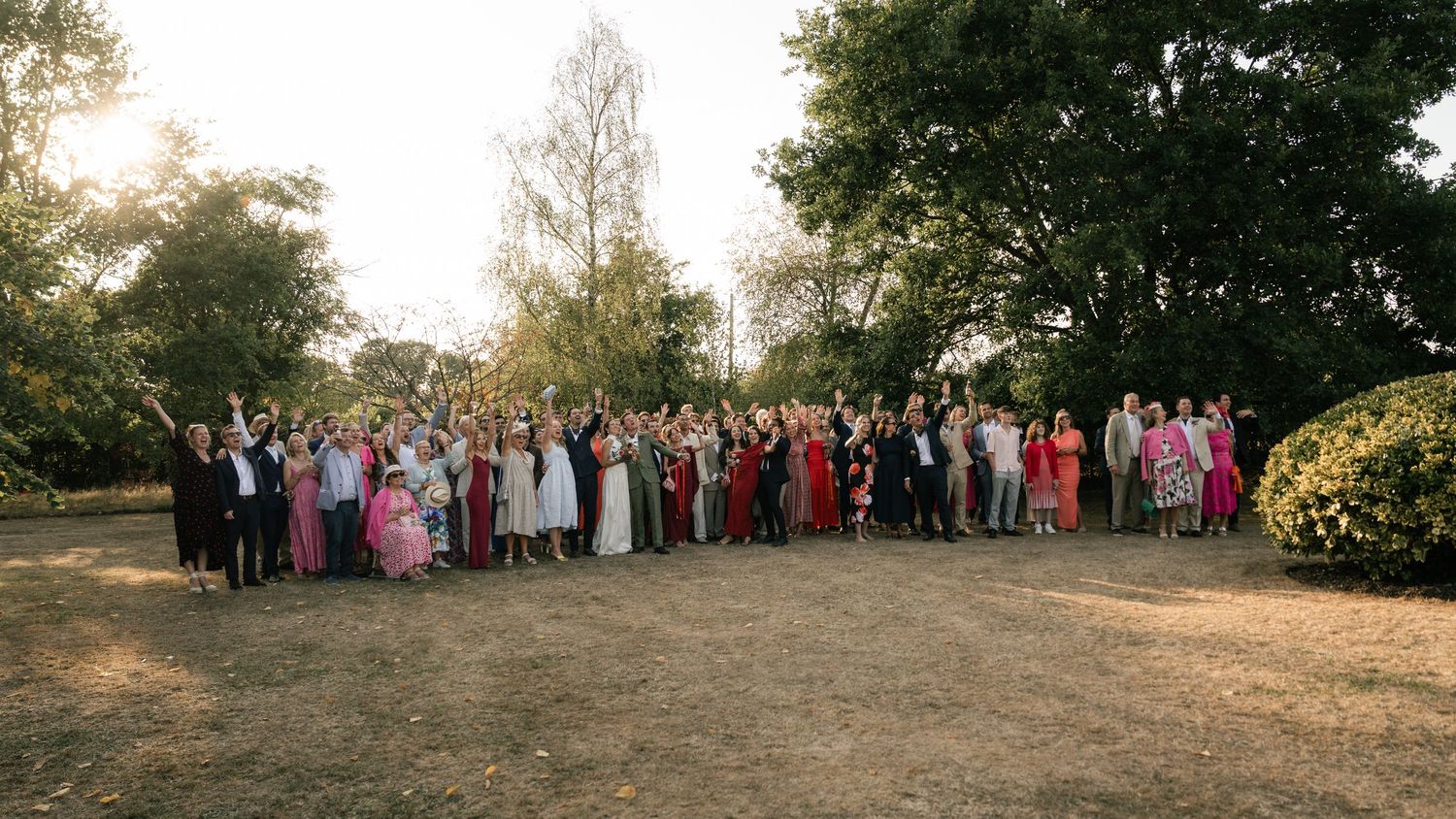 Large outdoor wedding party group photo with guests in formal attire gathered on a dirt path during golden hour.