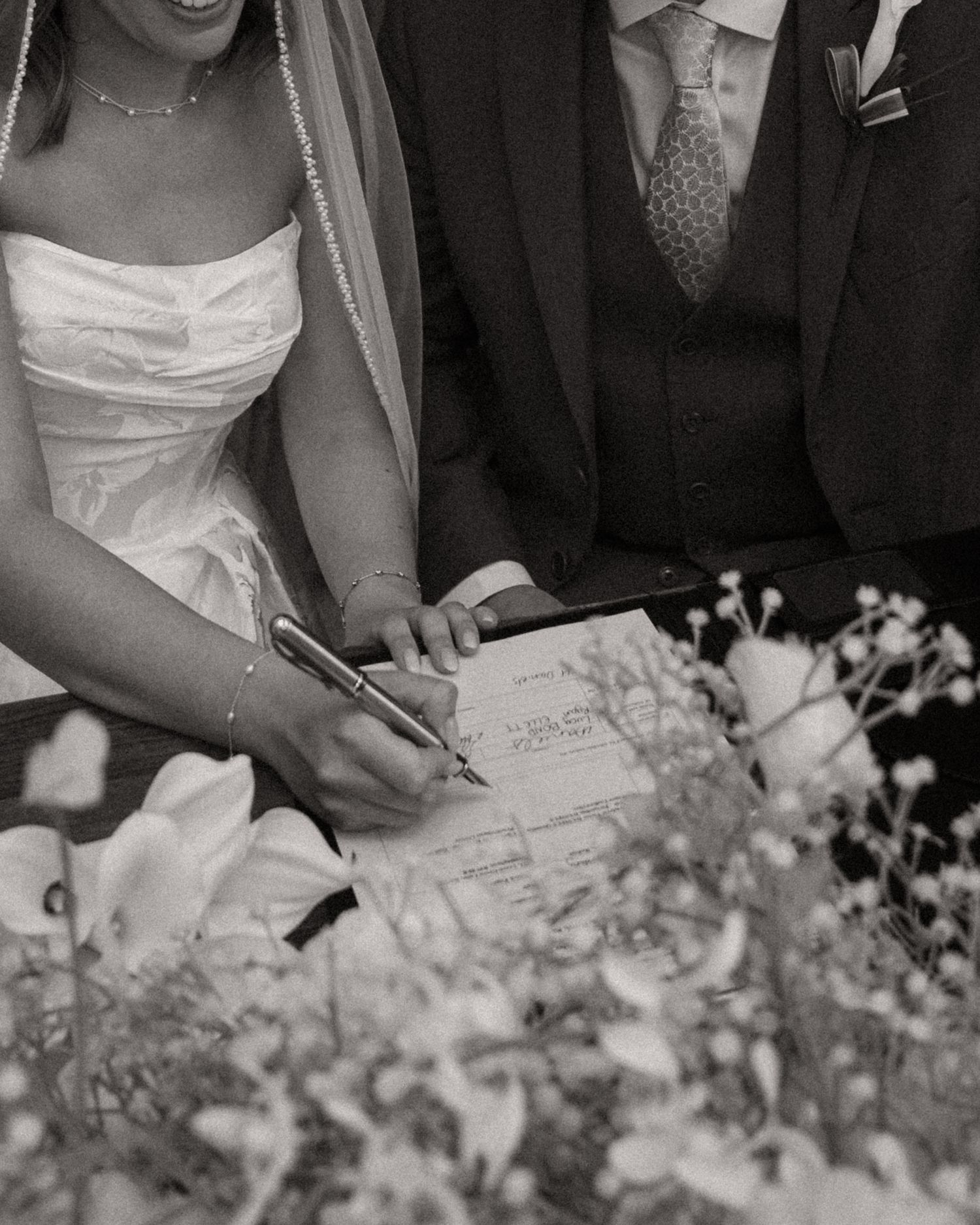 Black and white wedding photograph showing people signing documents with white flowers in the foreground.