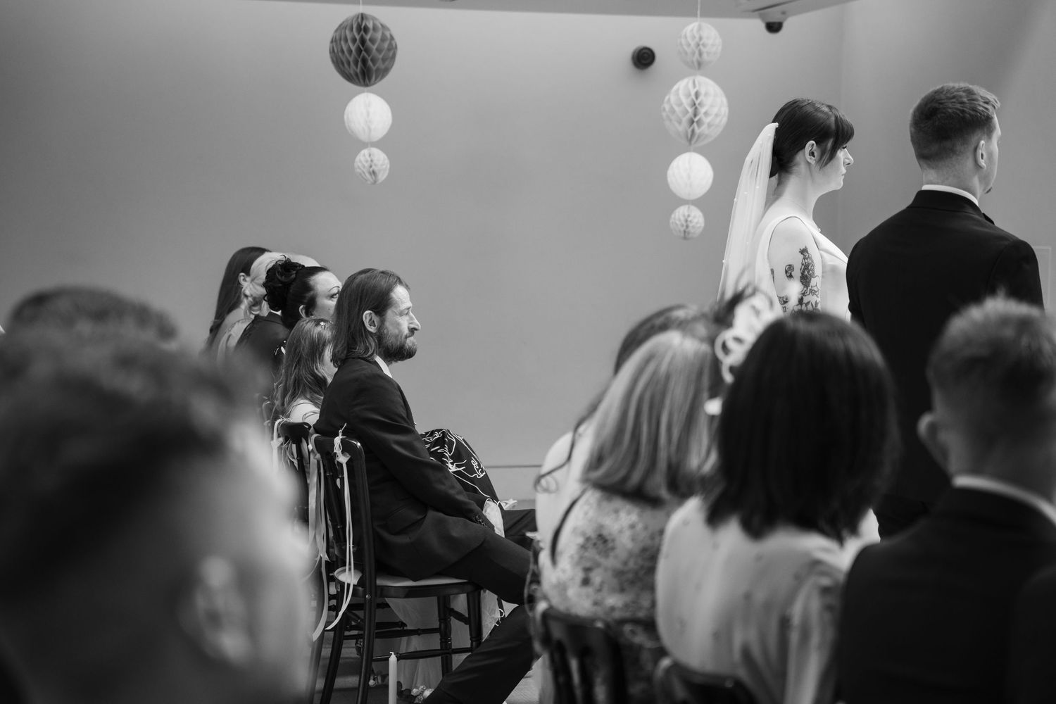 A black and white photograph of a wedding ceremony with guests seated beneath decorative hanging globe lights.