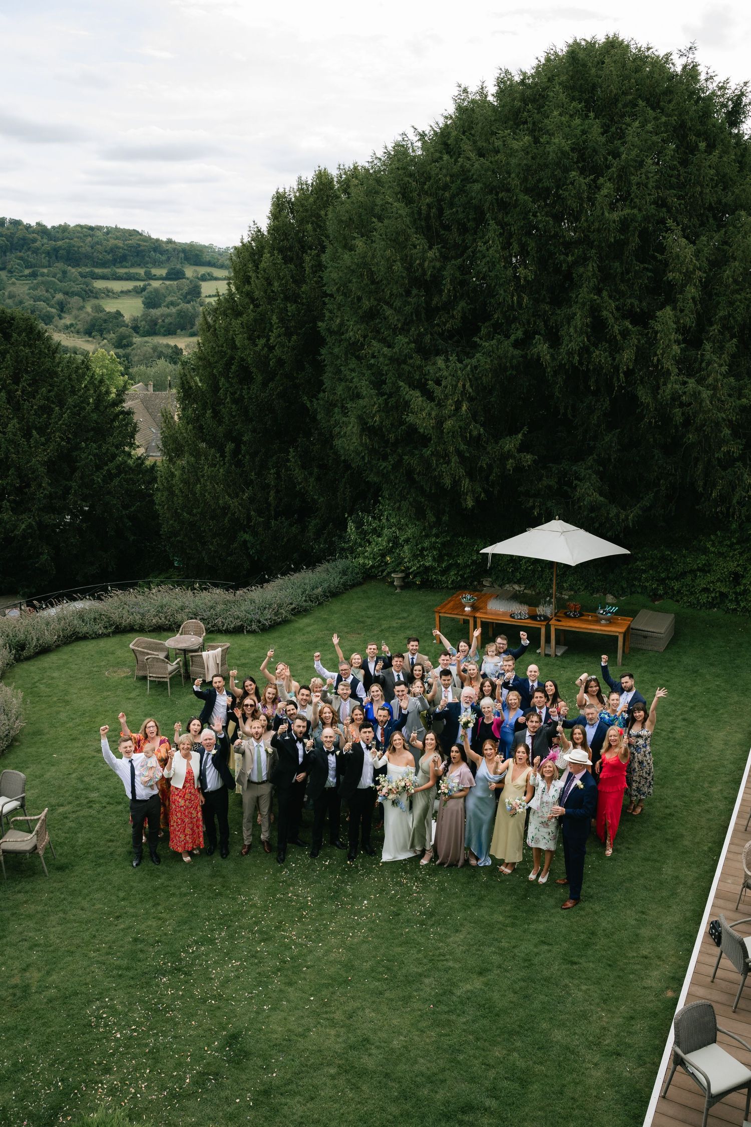 Bird's eye view of wedding guests arranged in formation on a hillside lawn surrounded by trees.