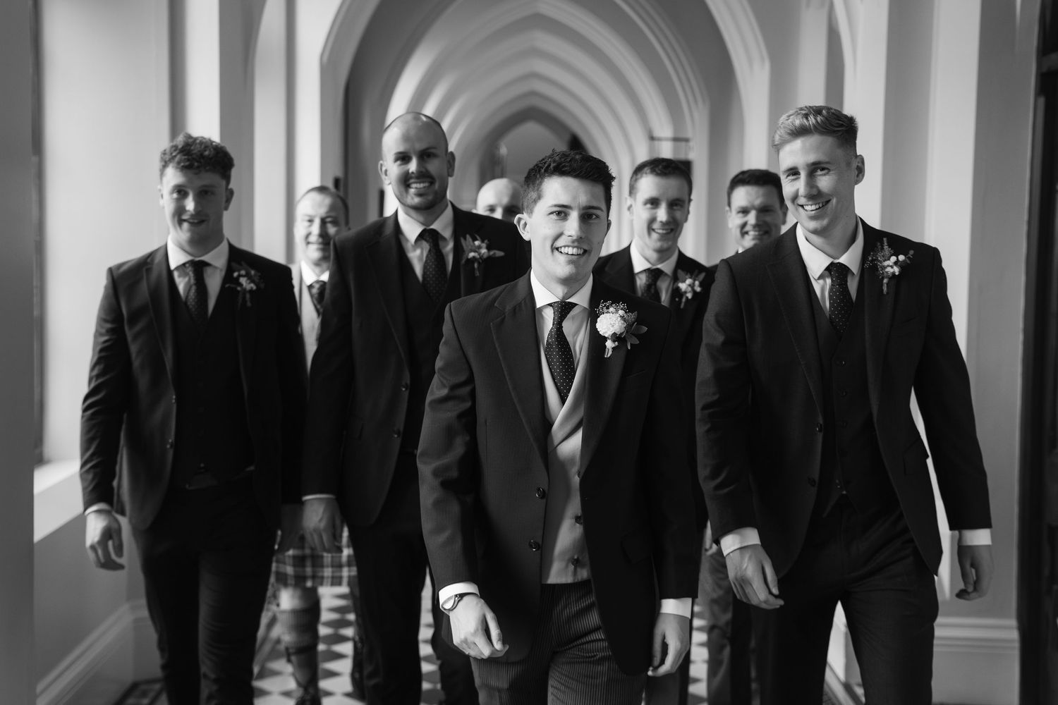 Groomsmen walking through arched hallway in formal suits.