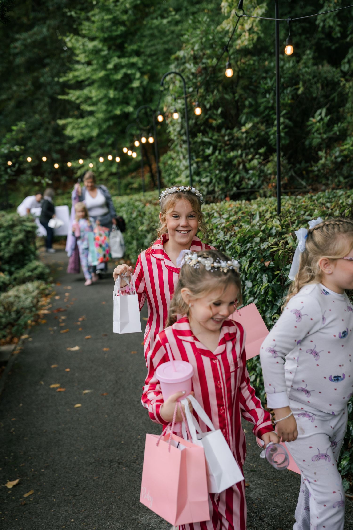 Children in pajamas and party dresses hold pink gift bags at an outdoor celebration with string lights.