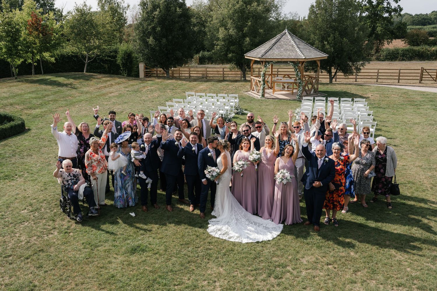 Wedding guests gather for outdoor group photos on a grassy lawn with a rustic gazebo in the background.