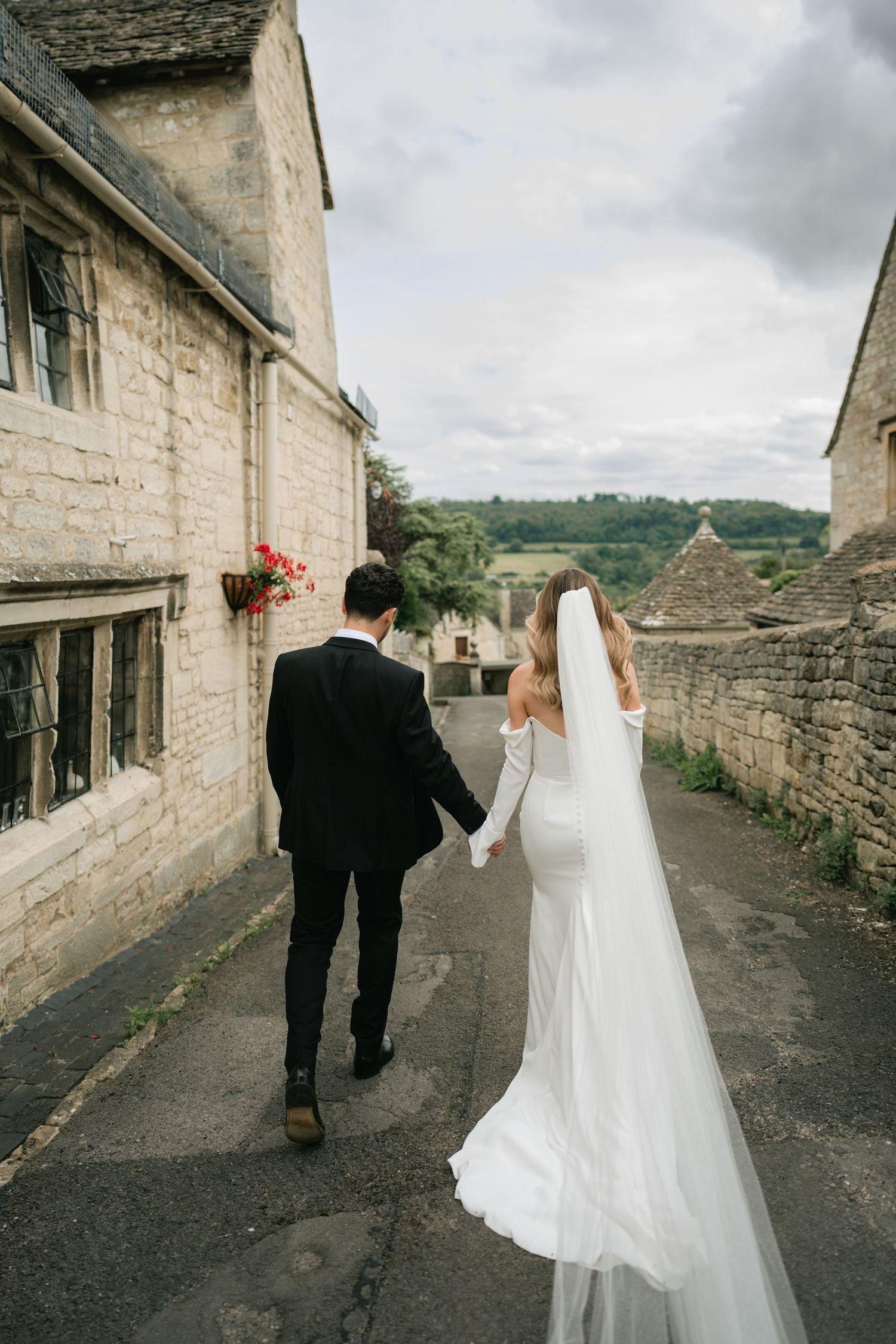 A couple in wedding attire walking down a stone-lined path in a rustic village setting under cloudy skies.