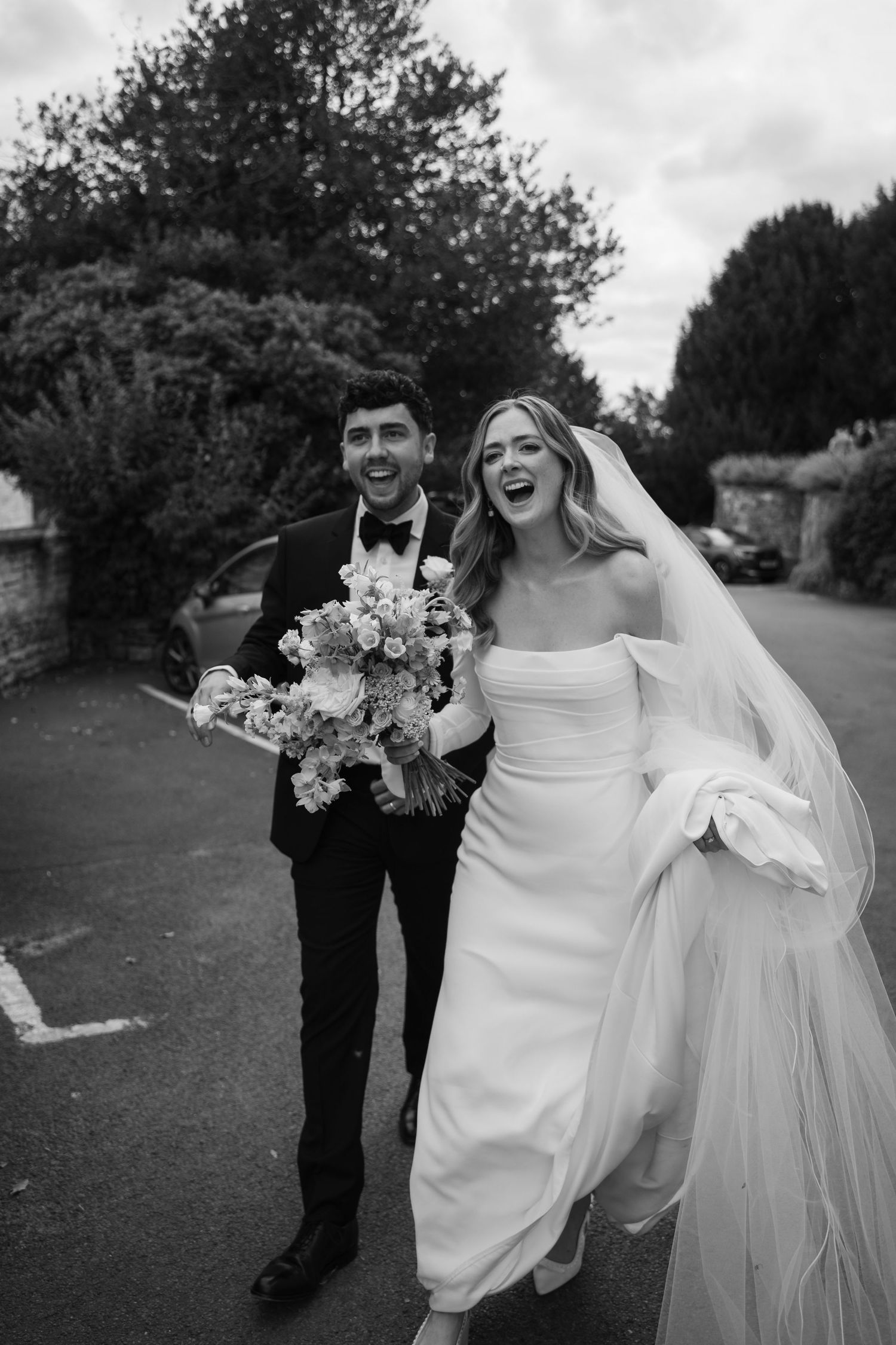 A joyful black and white wedding photo showing a couple laughing together while holding a floral bouquet outdoors.