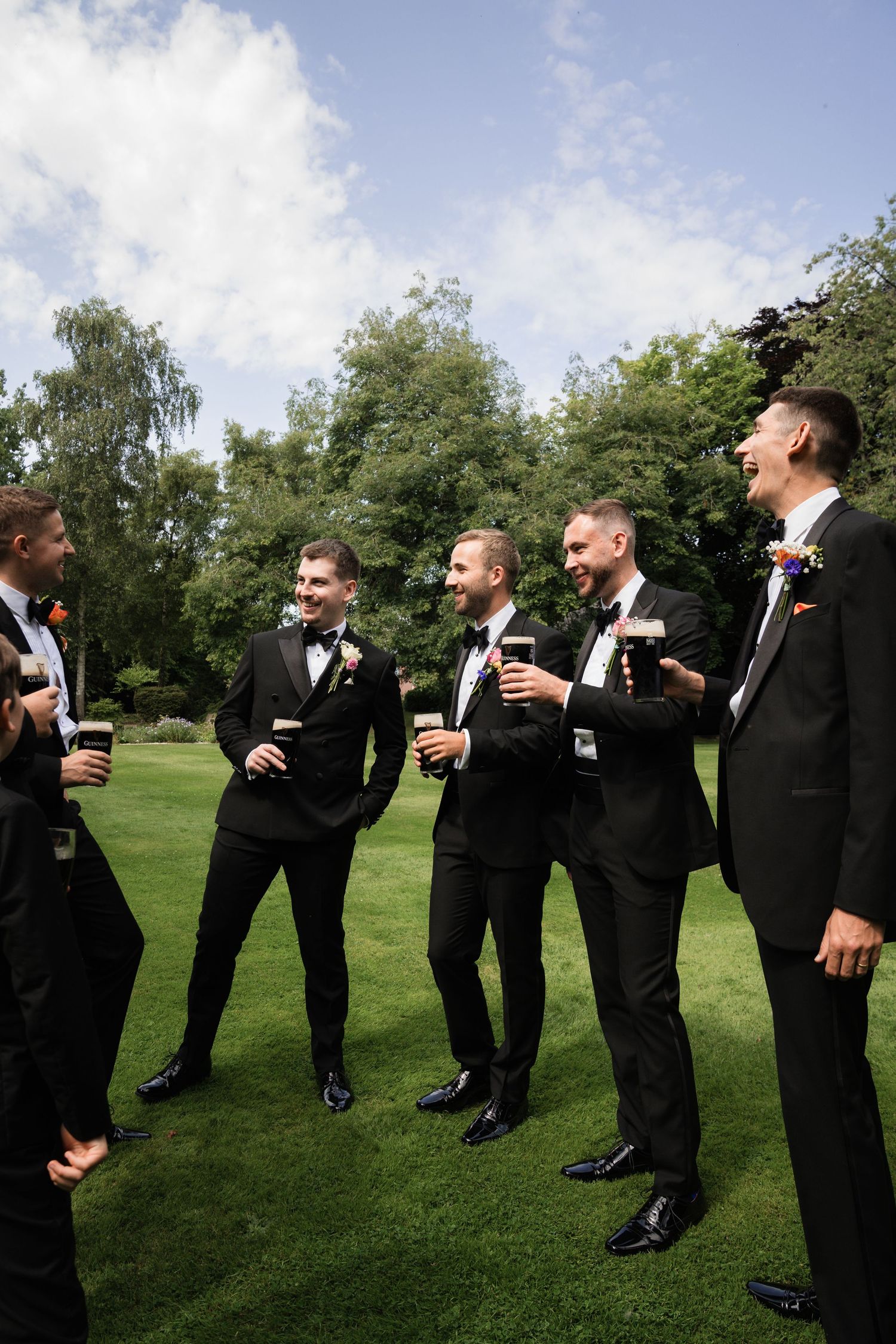 Groomsmen in black tuxedos stand in a line on green grass sharing drinks and laughs at an outdoor wedding.