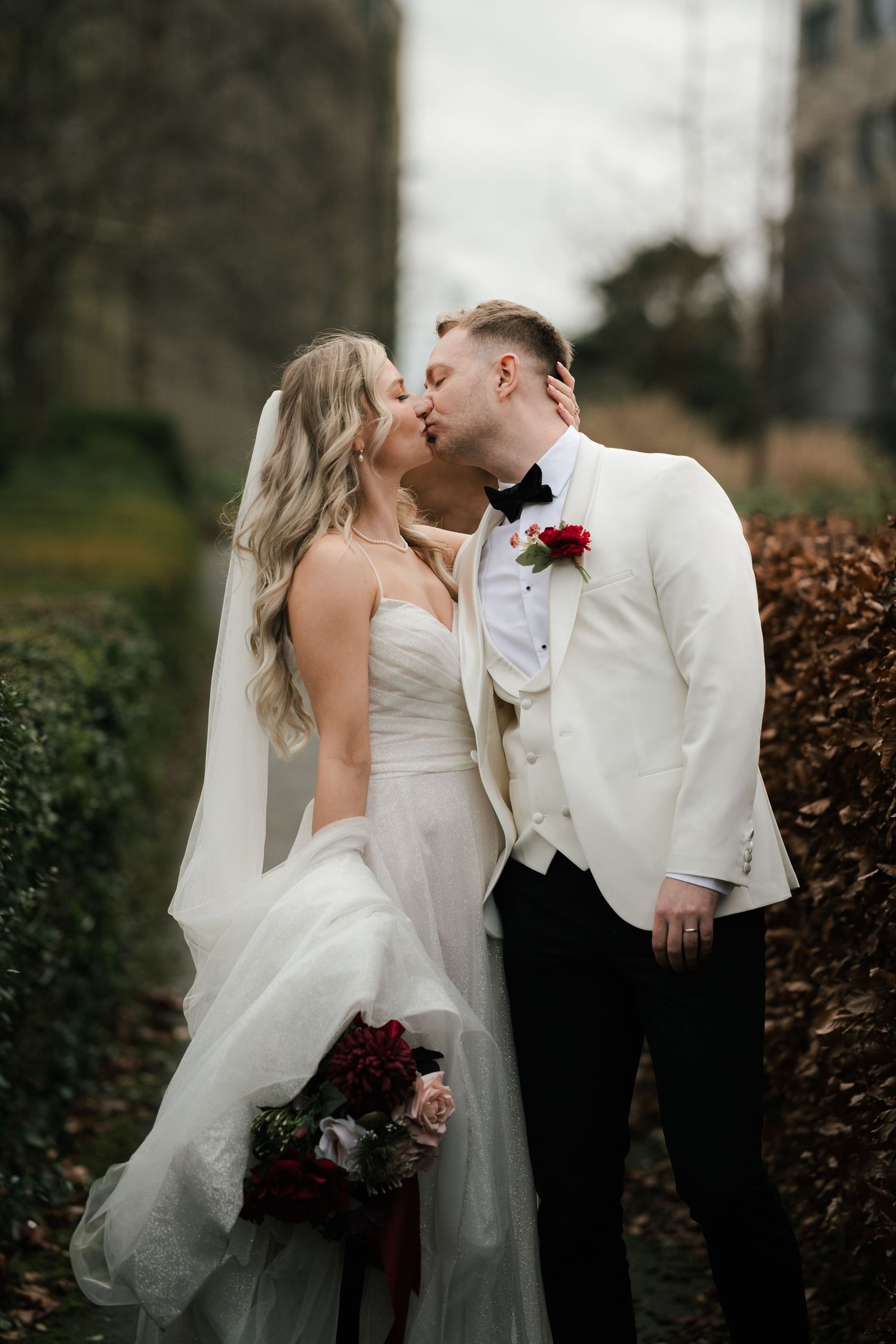 A romantic wedding couple shares an intimate kiss in a garden setting while holding a burgundy bouquet.