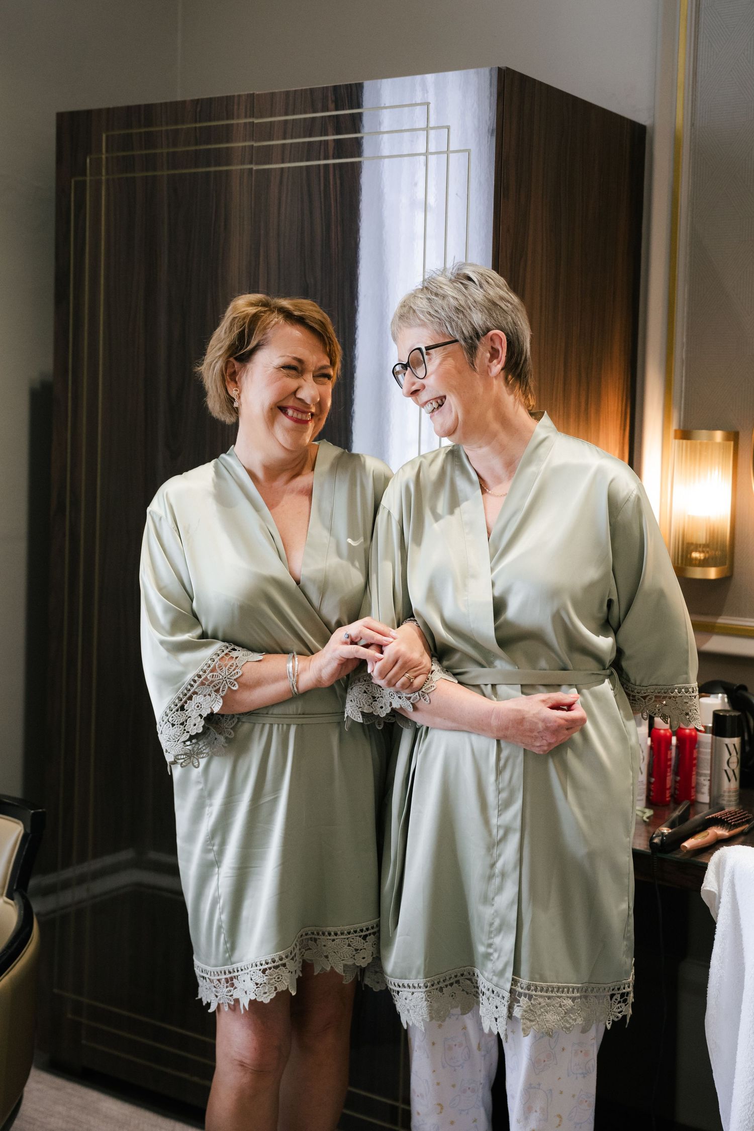 Two people in matching sage green robes share a tender moment while getting ready in an elegant room.
