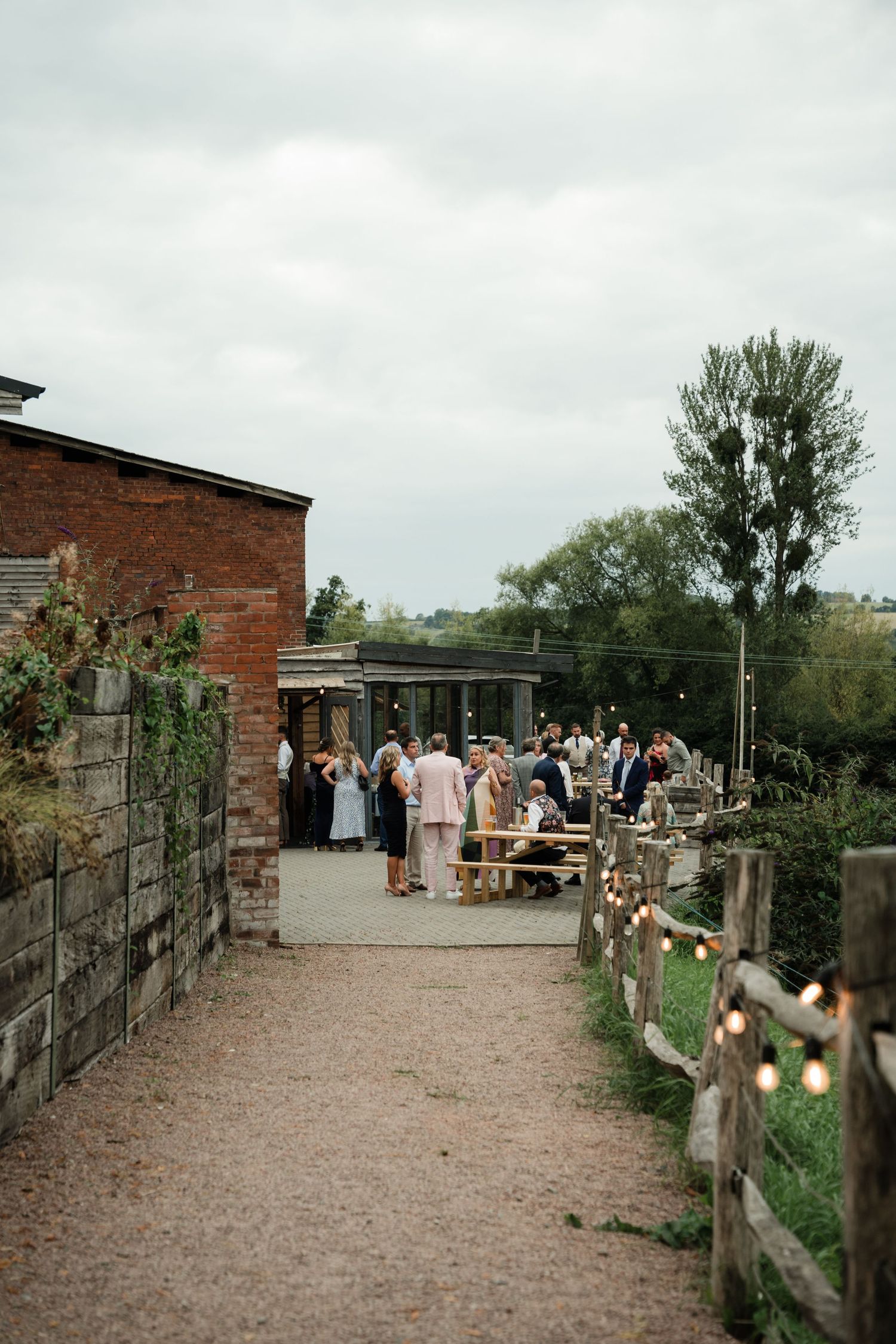 Rustic outdoor venue with brick buildings, wooden fences, and gravel paths where people gather for a social event.