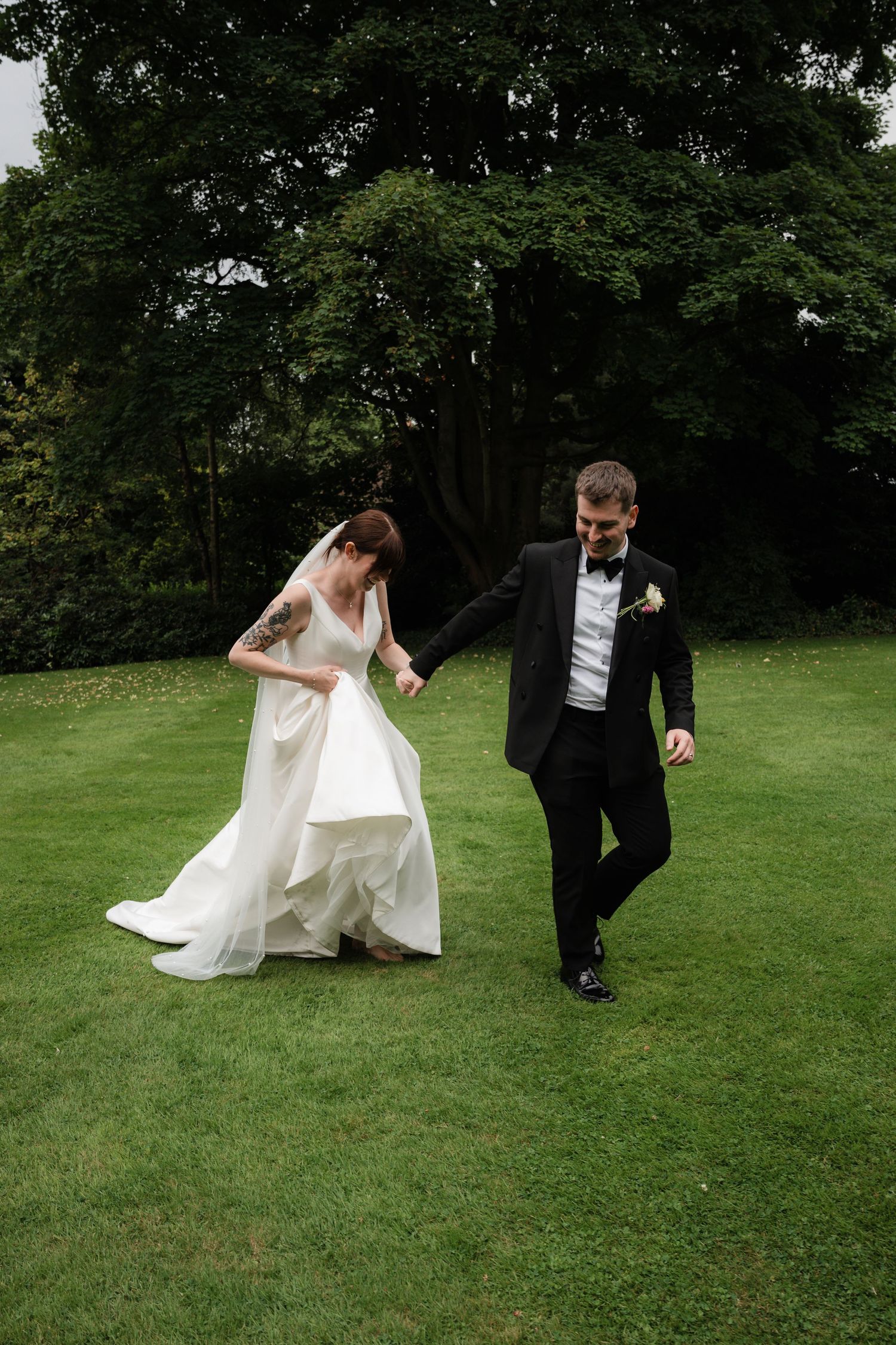A couple in wedding attire walks together on a lush green lawn surrounded by trees.
