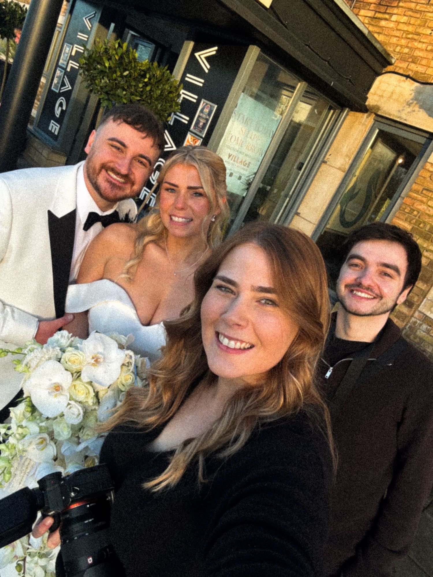 A group celebrates together at a wedding venue with a beautiful white floral bouquet and brick exterior.
