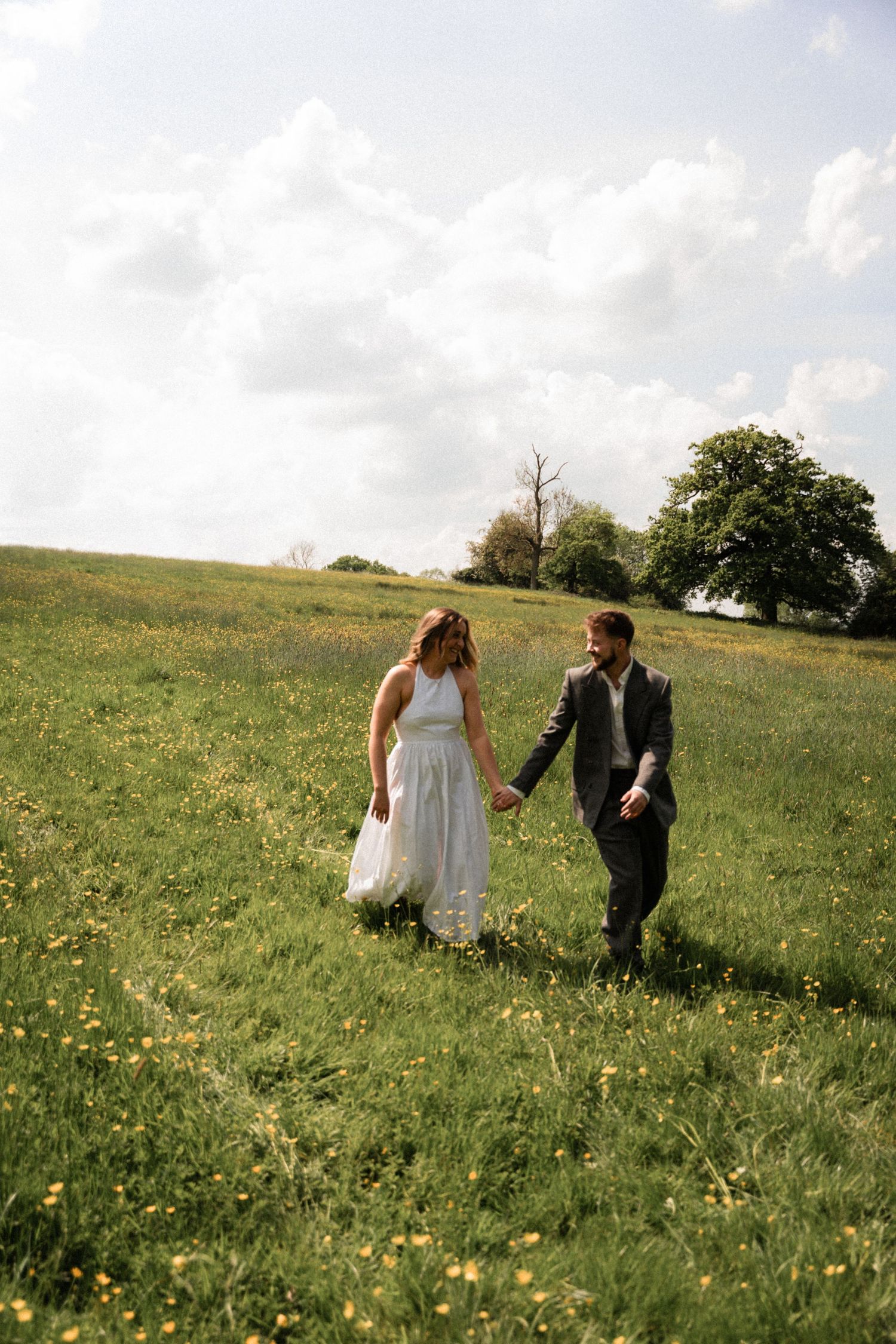 A couple walks hand in hand through a sunny meadow filled with yellow wildflowers.
