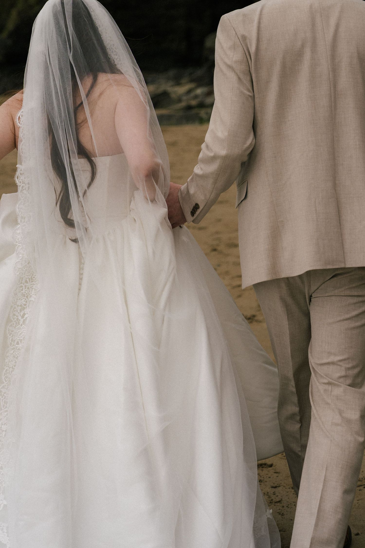 Bride in white gown and veil holds hands with groom in beige suit while walking on sandy beach.